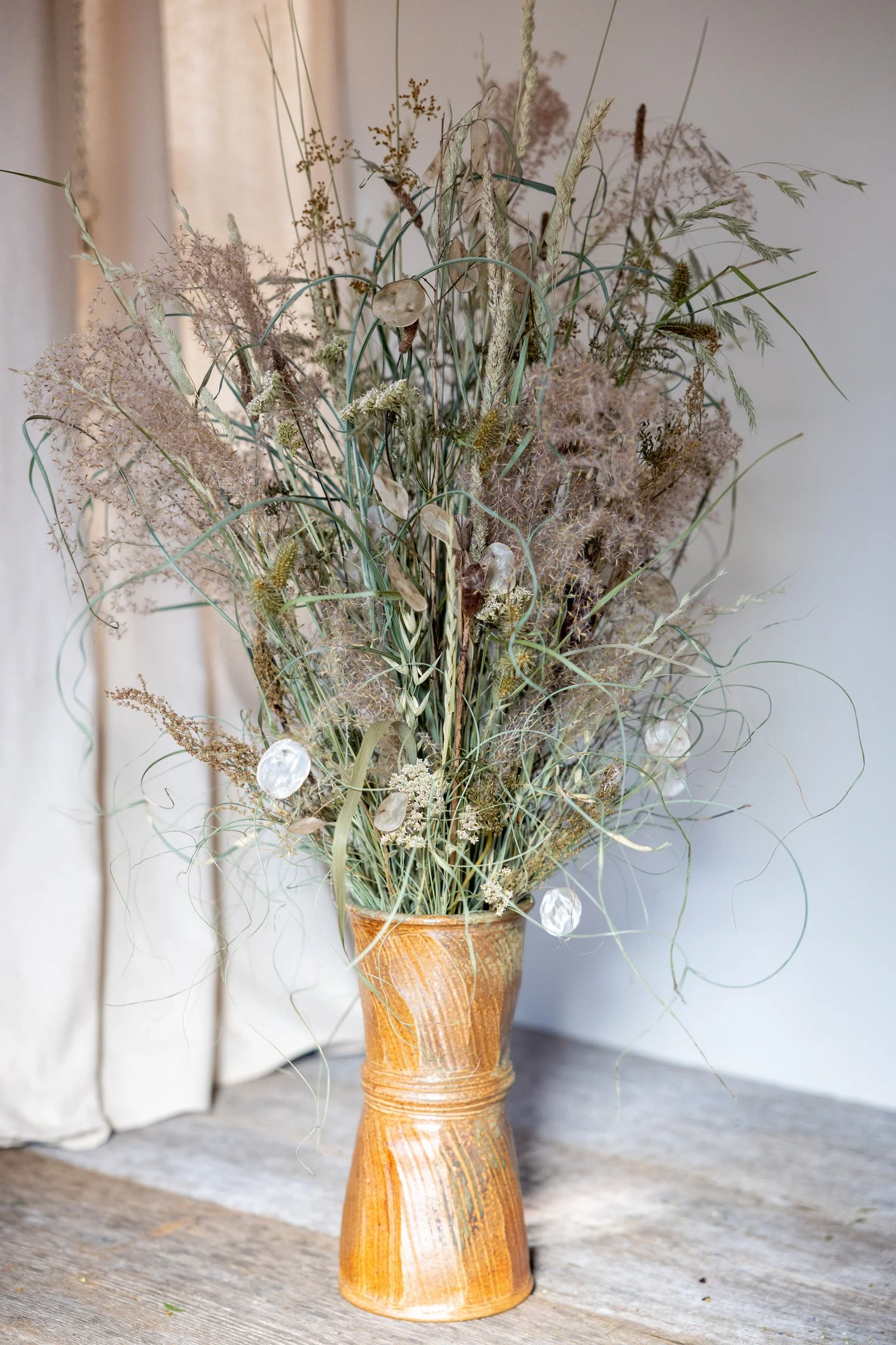 A rustic orange-brown ceramic vase with a narrow neck and wider base, filled with dried wildflowers and grasses, placed on a wooden surface against a neutral background.