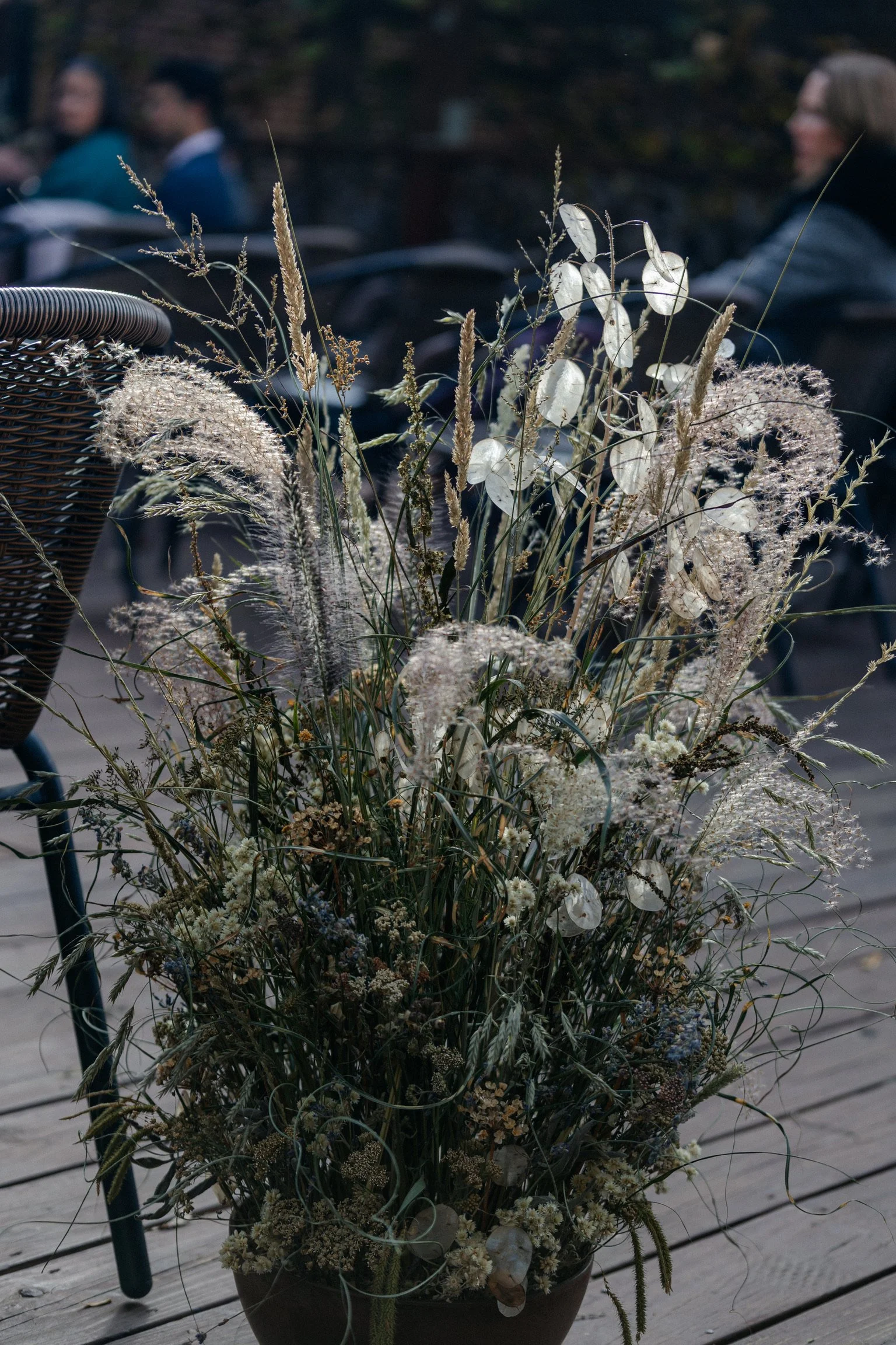 A large bouquet of dried flowers and grasses in a brown pot on a wooden deck, with people sitting and socializing in the background.