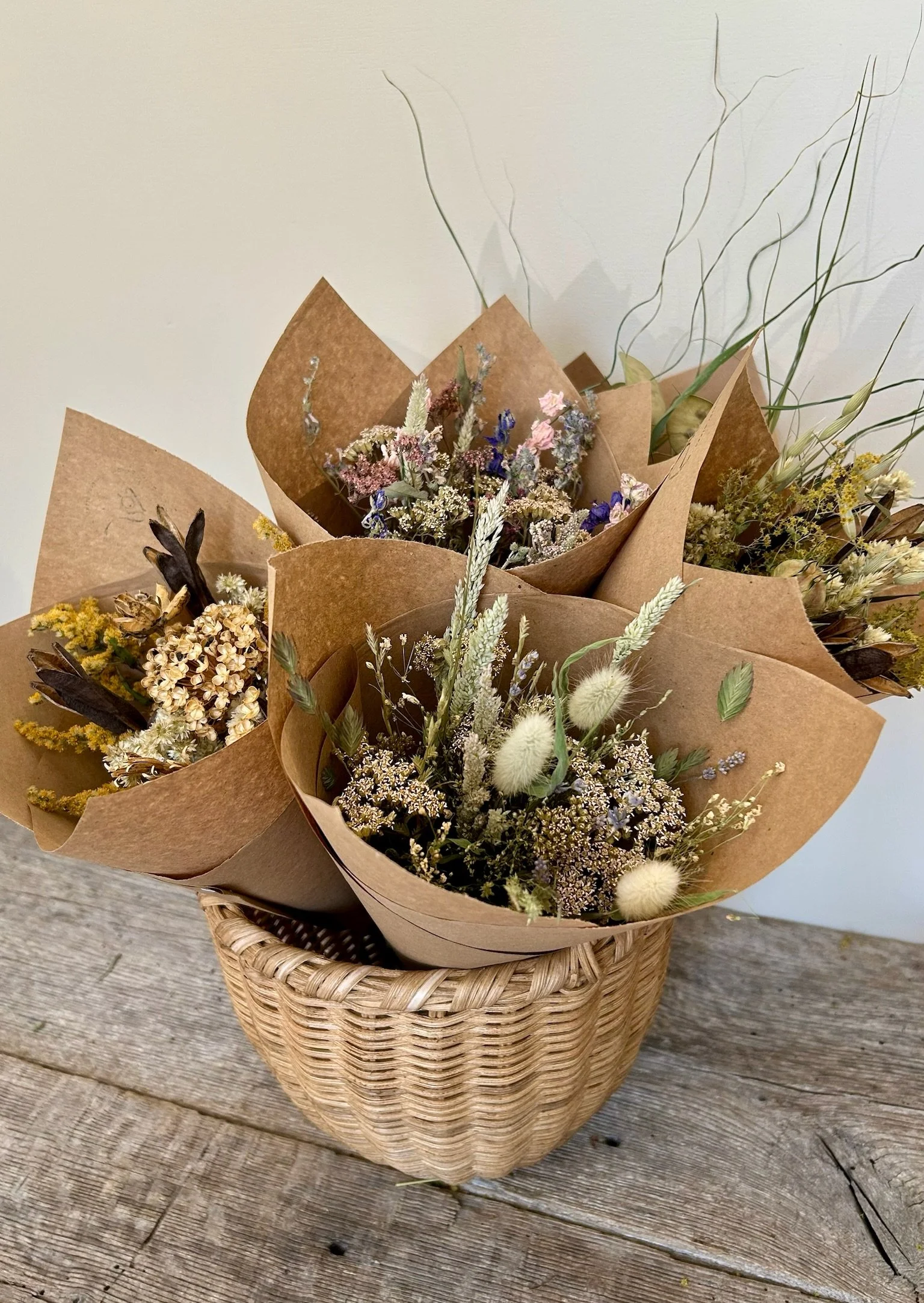 A wicker basket filled with bouquets of dried flowers wrapped in brown paper, placed on a wooden surface.