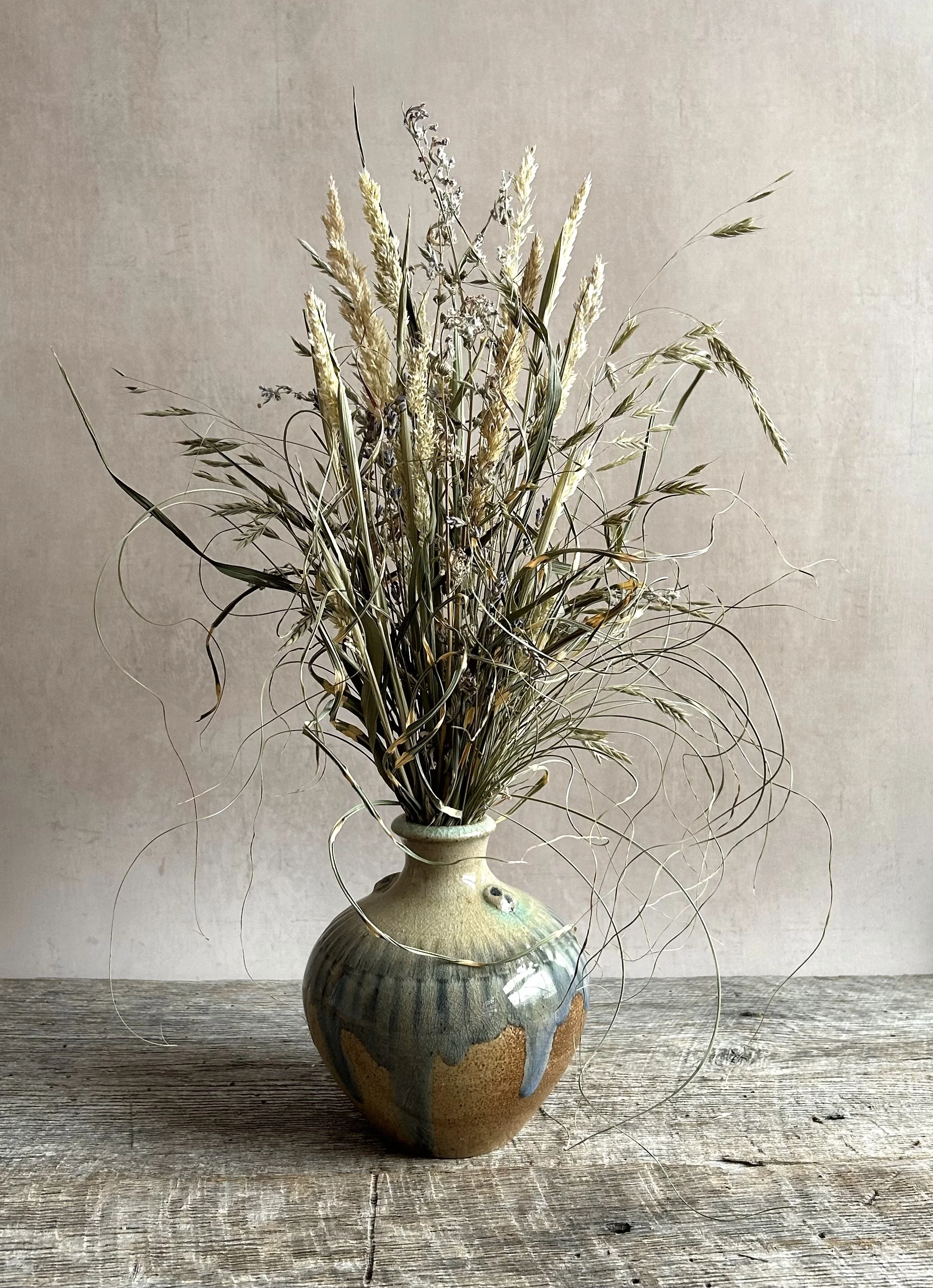 A ceramic vase with a rustic glaze, containing a dried flower arrangement with grasses and seed heads, set on a weathered wooden surface against a plain wall.