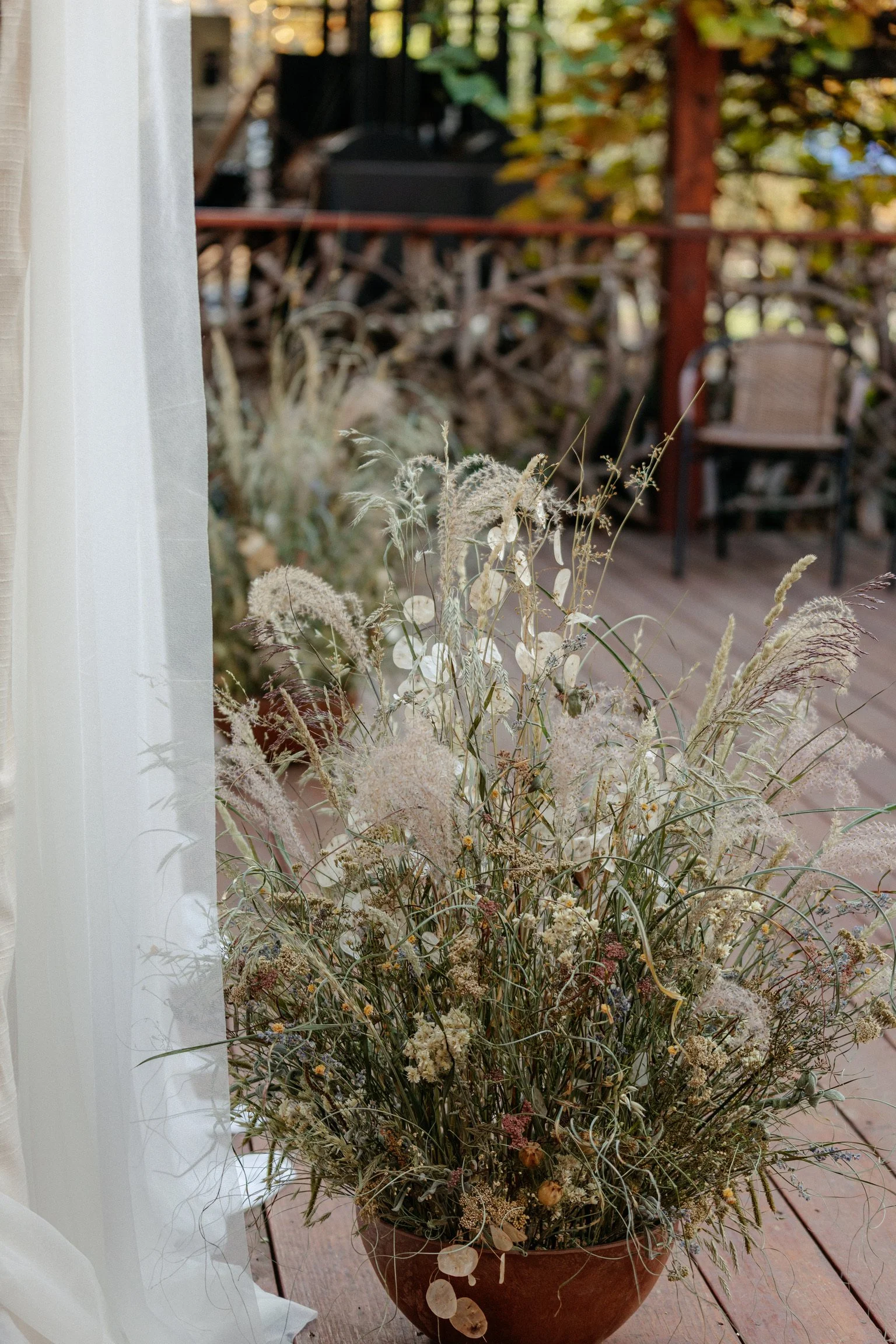 A potted arrangement of dried grasses and flowers on a wooden floor near a window with sheer curtains, outside a deck with chairs and plants.