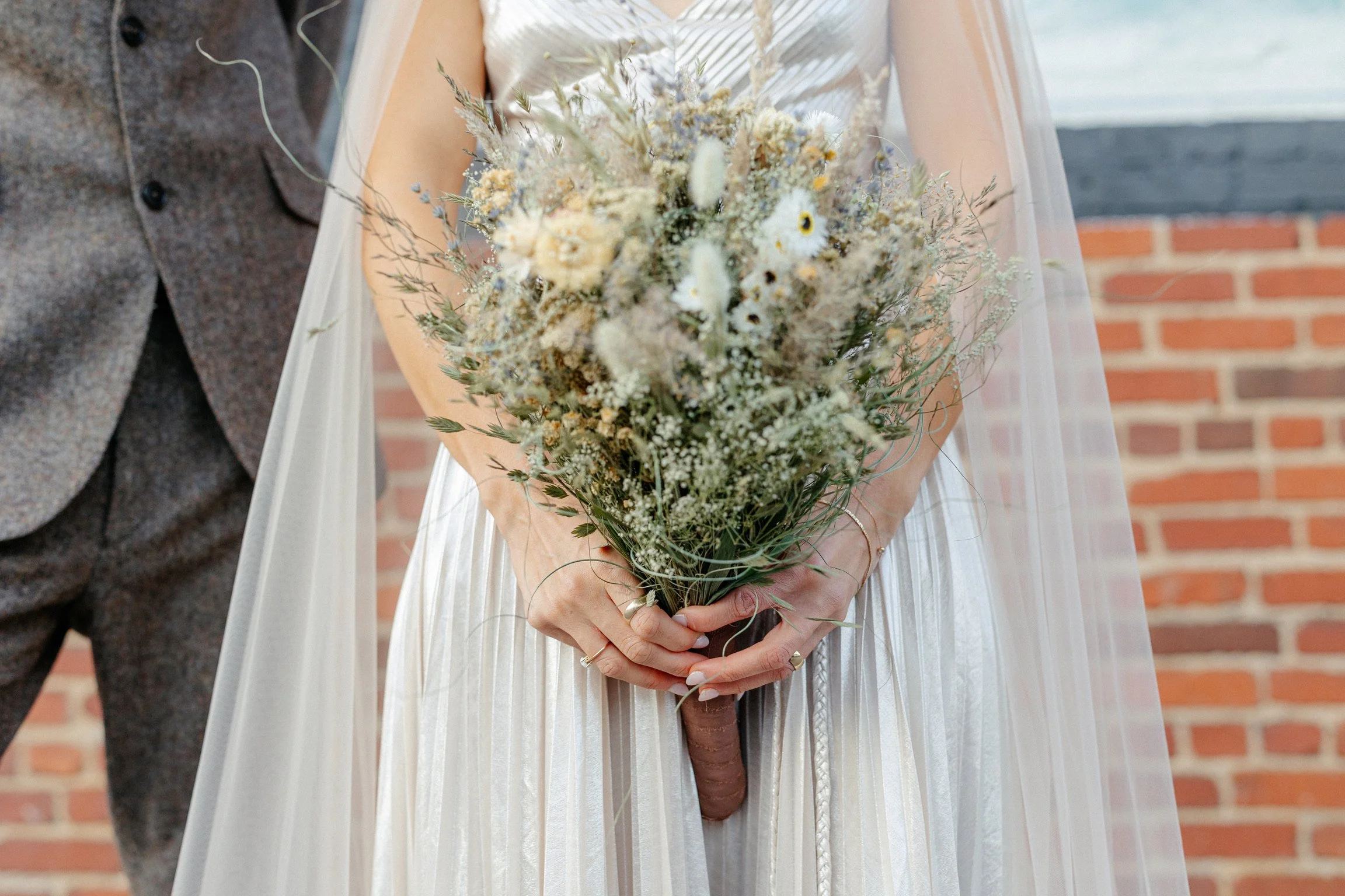 Bride holding a bouquet of flowers in front of her waist, with a groom partially visible on the left, against a brick wall background.