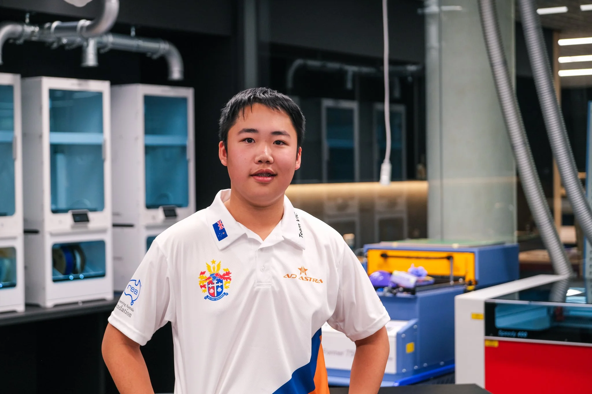 Young man in a white polo shirt with Australia and team logos standing in a modern industrial or research facility with various equipment and machinery in the background.