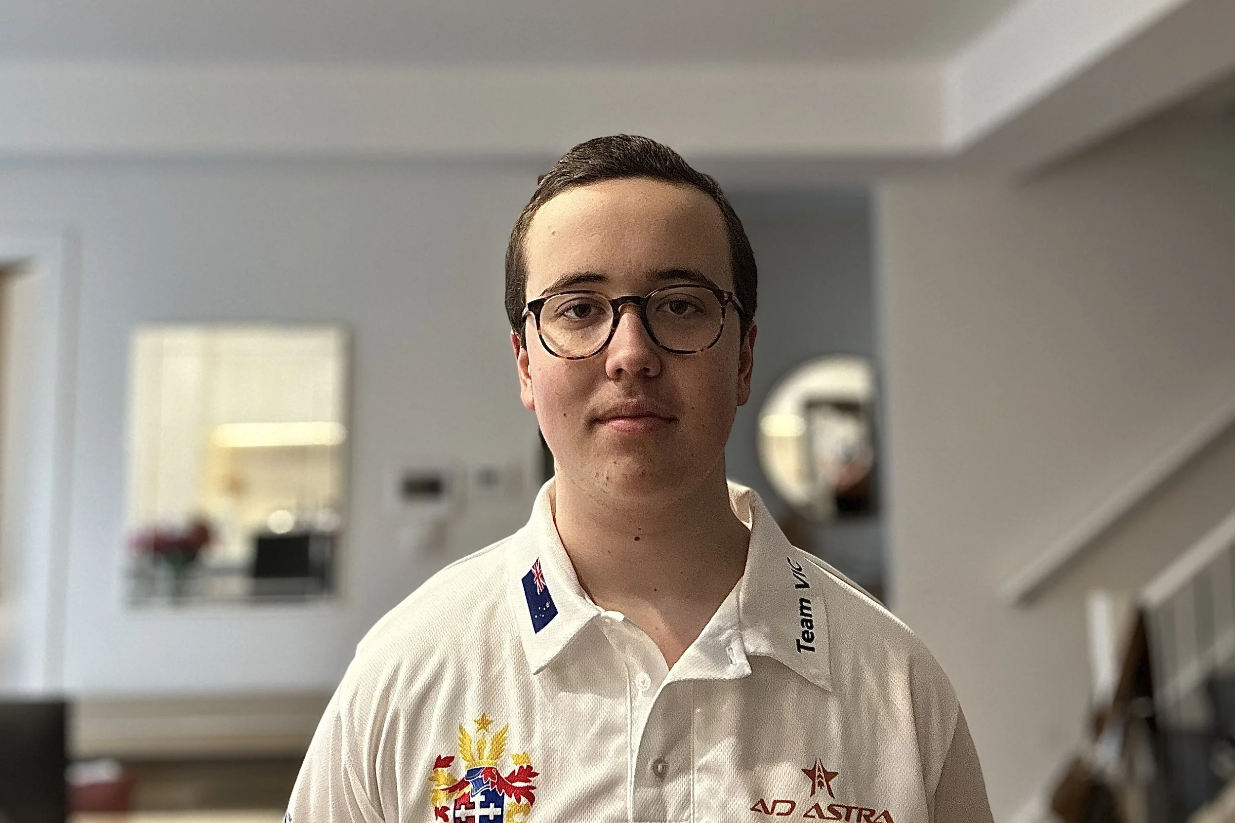 Young man with glasses wearing a white sports polo shirt with logos and flags, standing indoors in a modern home.