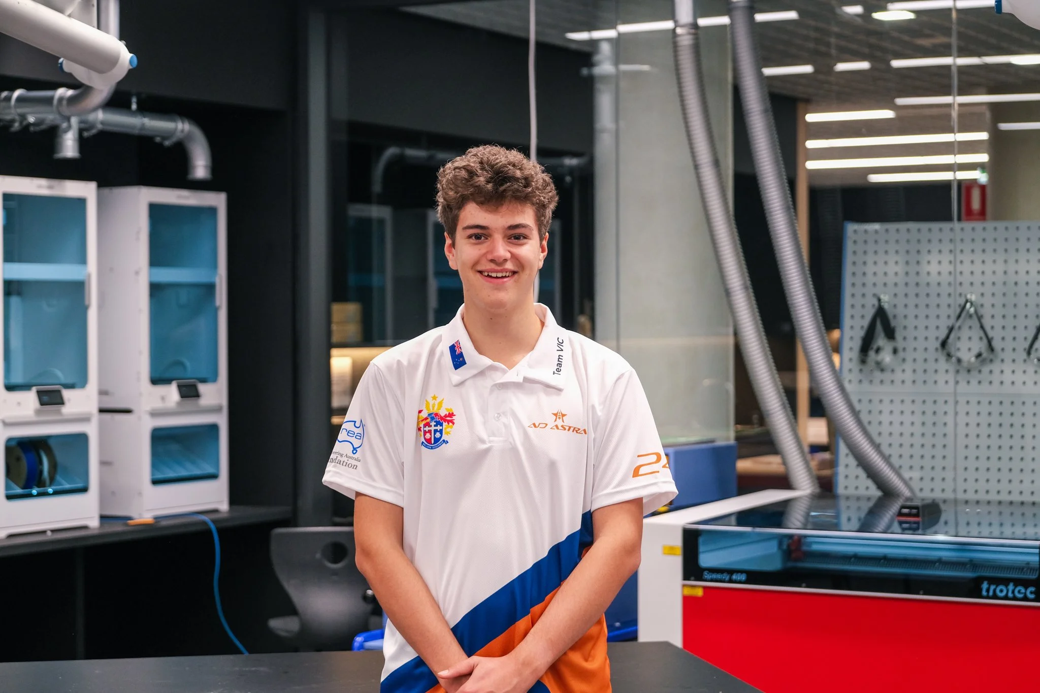 A young man with curly hair smiling in an indoor laboratory or workshop setting, wearing a white polo shirt with various logos and team branding.