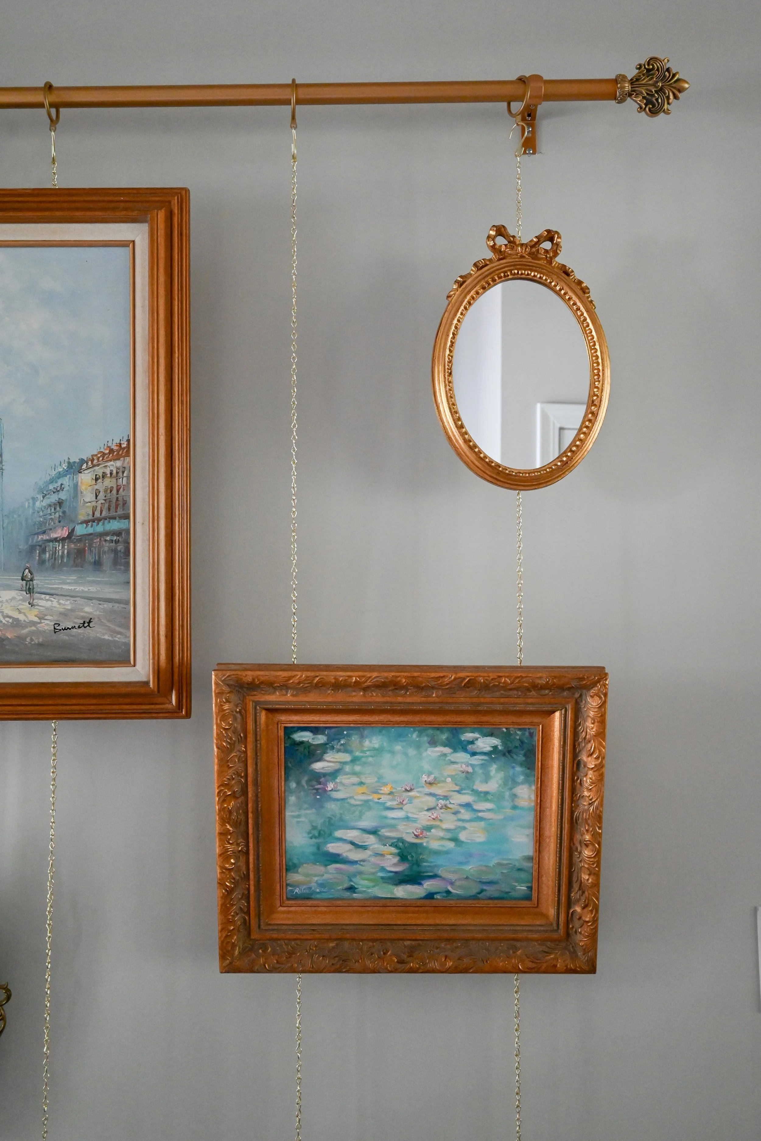 Gallery wall styled with a picture rail and antique brass hooks, featuring a signed oil painting of a Parisian street scene, a small oval gilt bow-top mirror, and a framed water lilies oil painting in an ornate gilt frame against a grey-green wall.