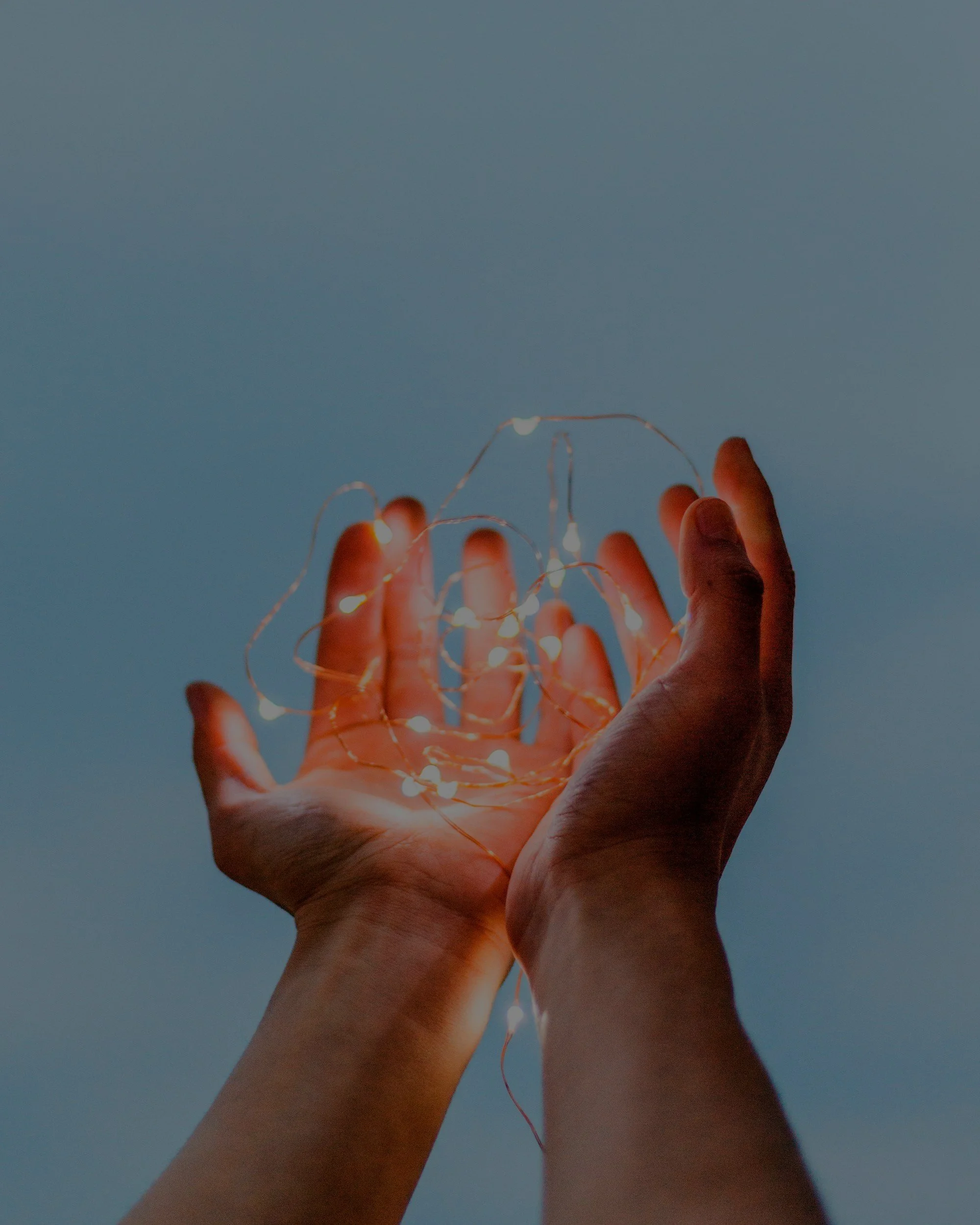 A pair of hands holding a tangled string of small warm white fairy lights against a light blue background.