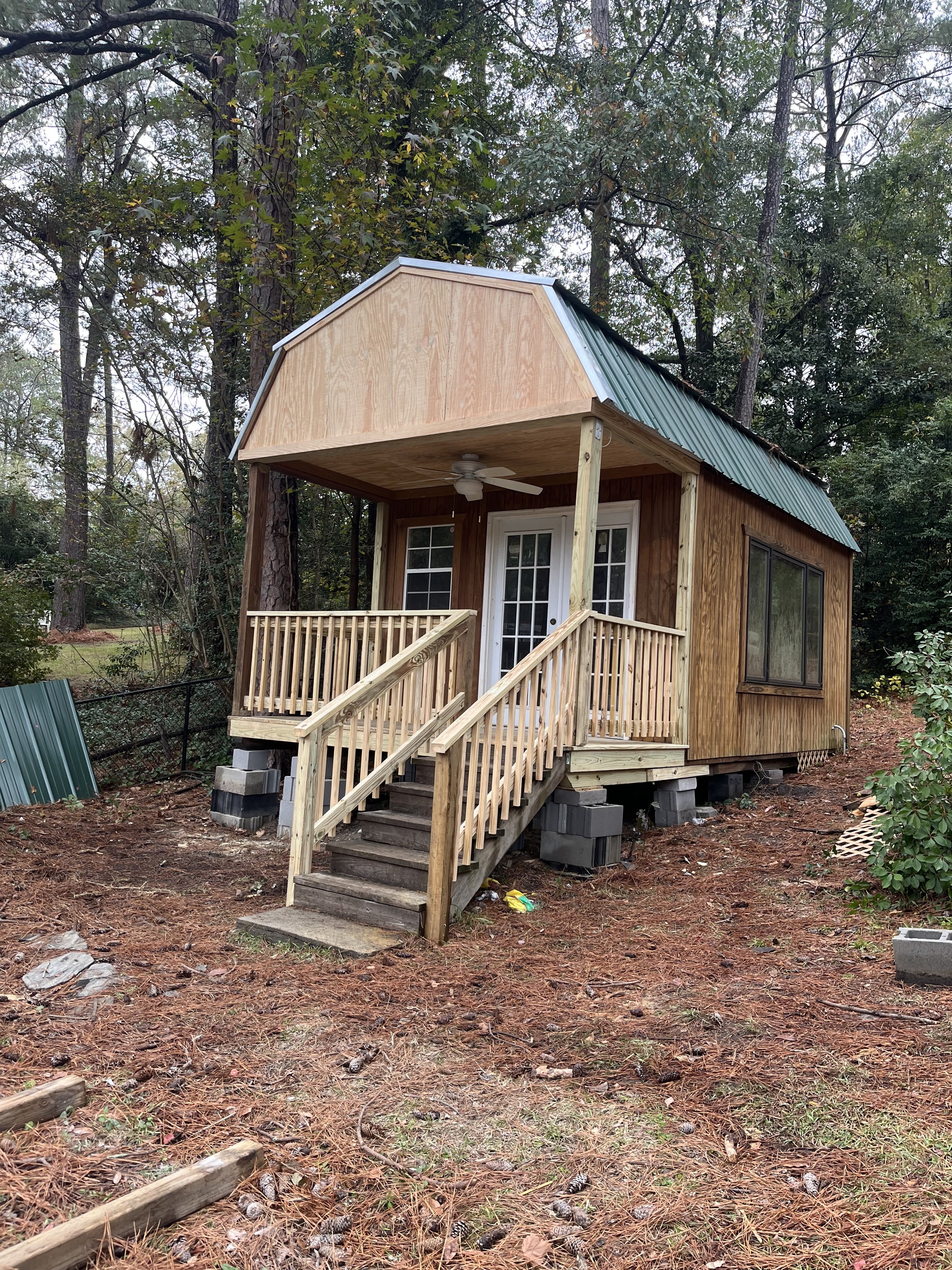 Small wooden house under construction with stairs and a porch, surrounded by trees and dirt ground.