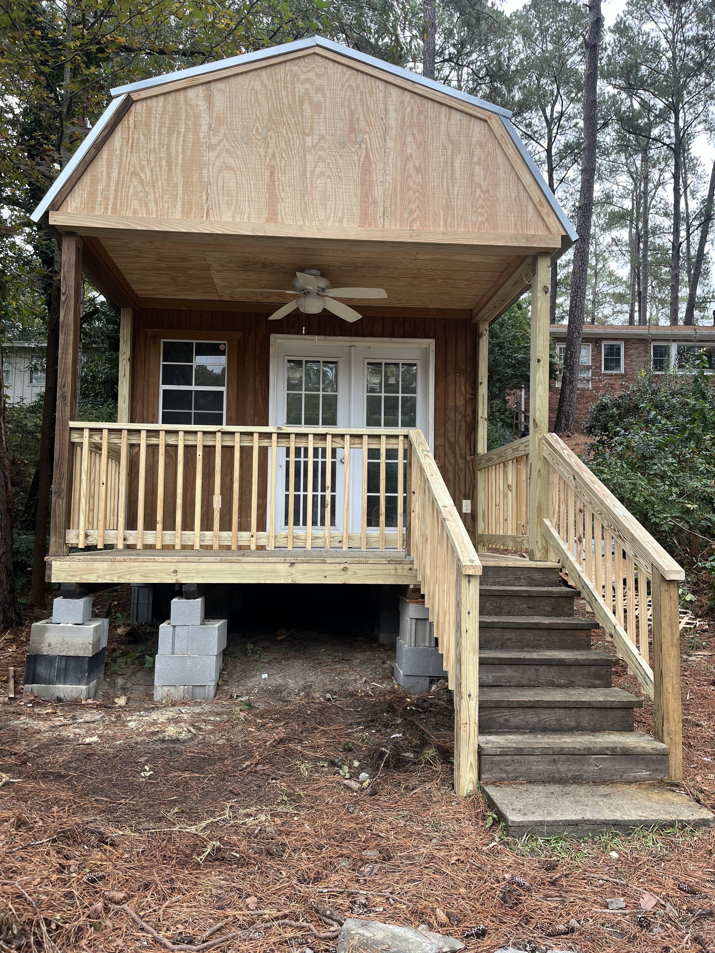 Small house with a wooden porch, white double doors, a ceiling fan, and partial stairs, in a wooded area.