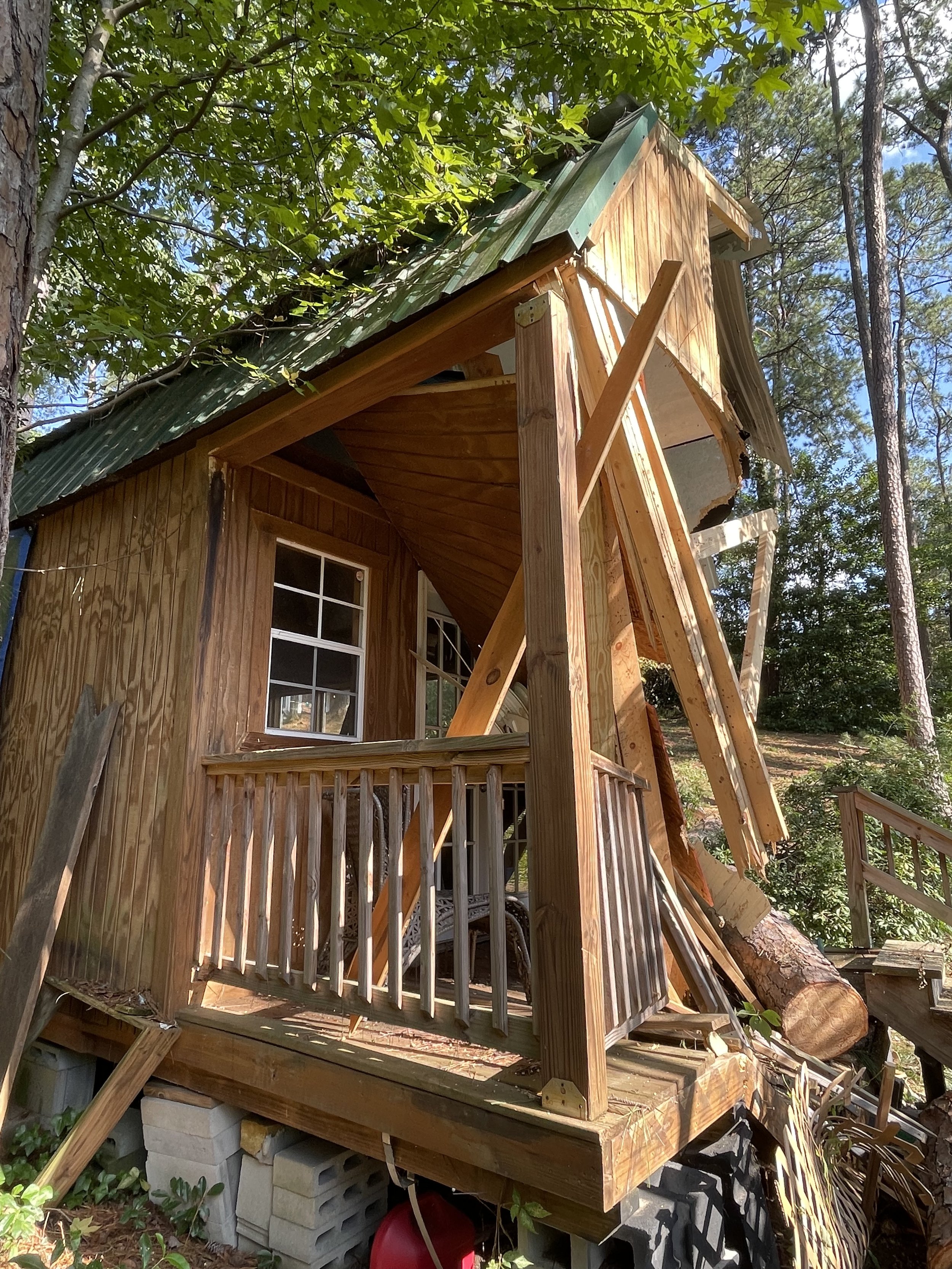 A partially constructed small wooden house with a green metal roof, surrounded by trees, with visible support beams and construction materials.
