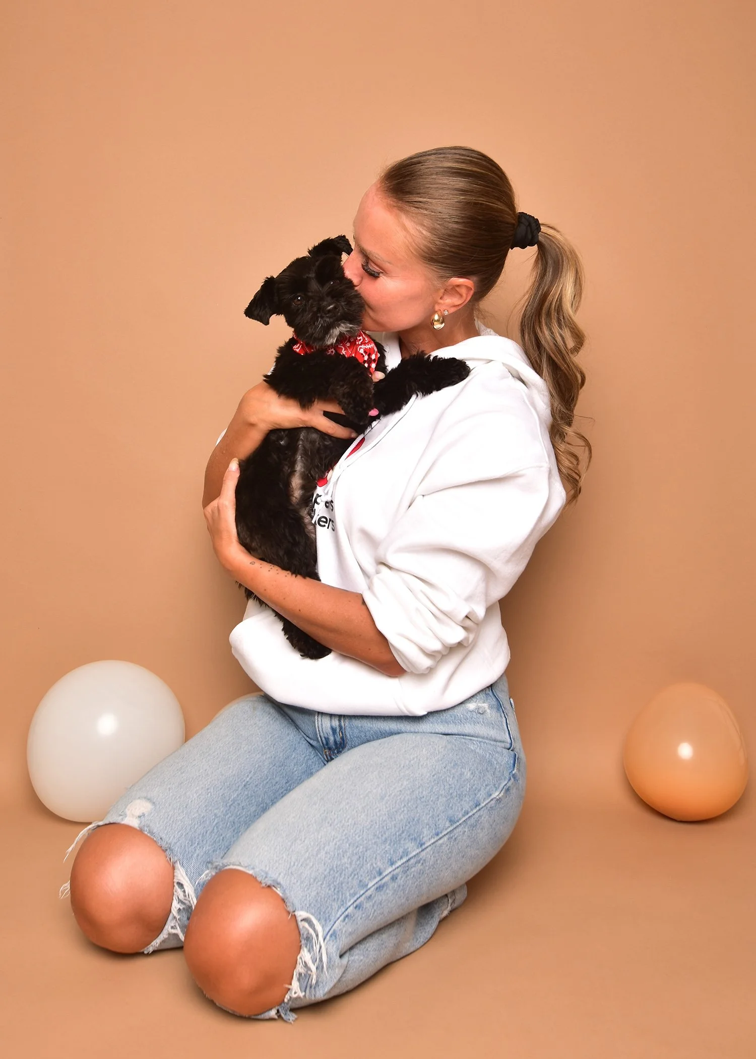 A woman with light brown hair tied back in a ponytail is kneeling and holding a small black puppy with a red bandana, giving it a kiss on the head. The background is beige with two balloons, one white and one peach, placed on the ground behind her.