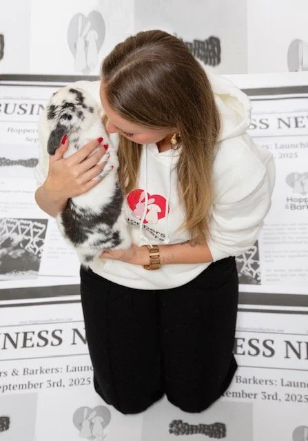 A woman holding a small, black and white puppy close to her face, with the puppy looking up at her, in front of a newspaper display