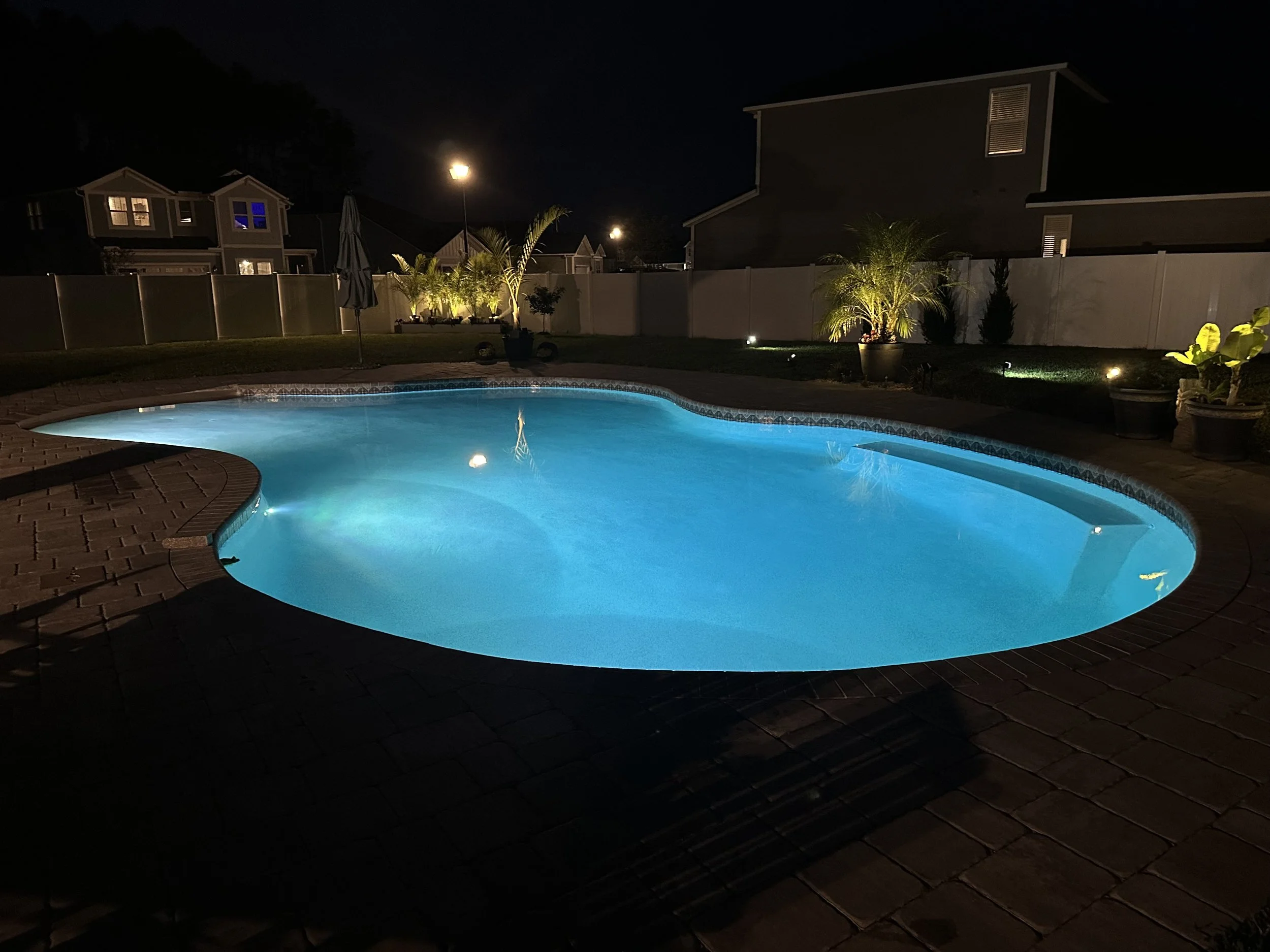 Night scene of a lit swimming pool in a backyard, surrounded by potted plants and outdoor lighting, with houses and a fence in the background.