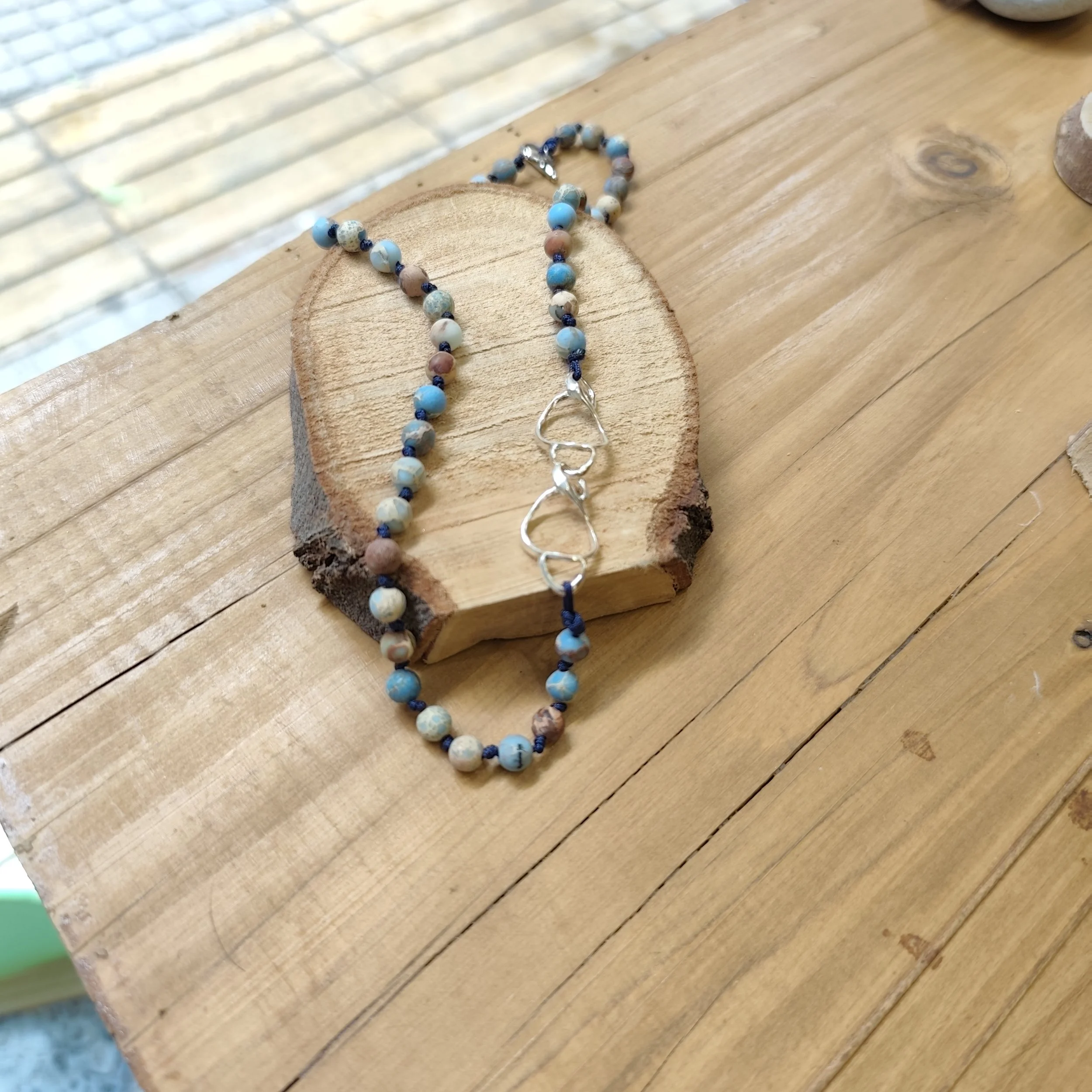 A beaded necklace with silver chain shape on a wooden surface with a small piece of wood underneath.