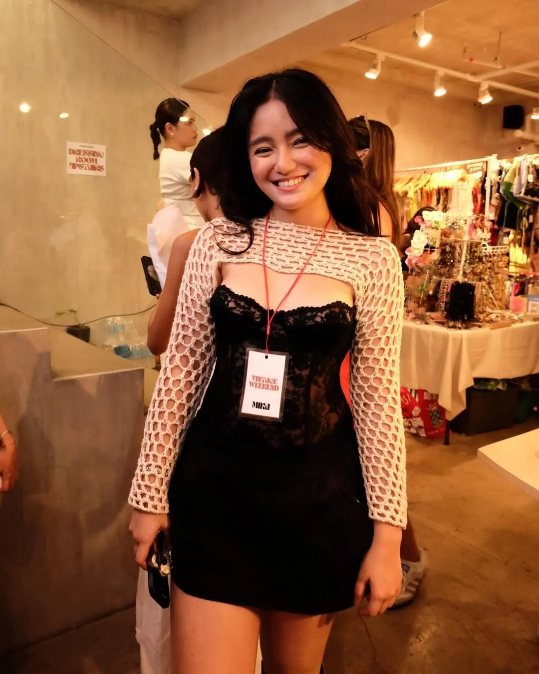 A young woman in a black lace dress with a beige netted cover-up, smiling at a fashion event with a name tag that reads 'VINTAGE WEEKEND MIKA.' She is in an indoor setting with booths and other attendees in the background.