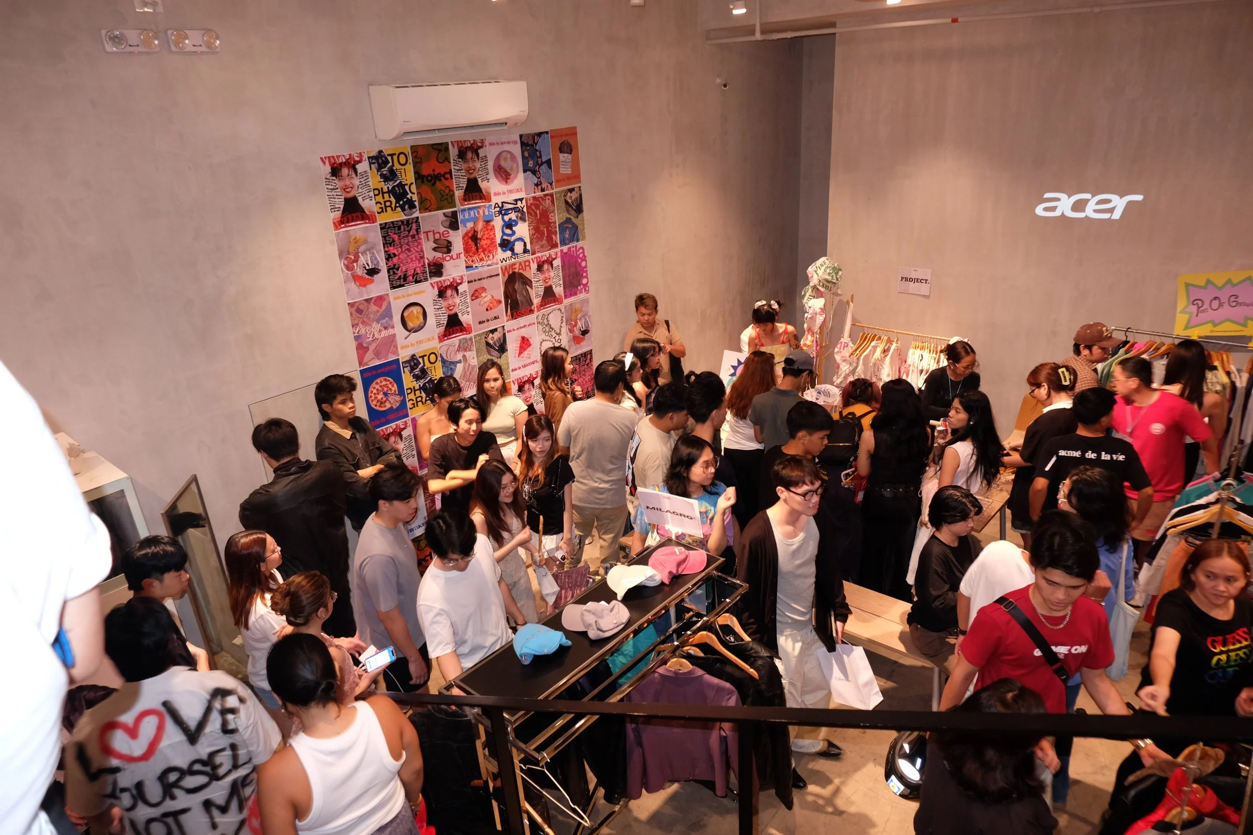 Crowd of people shopping inside a clothing store with racks of clothes and colorful posters on the wall.