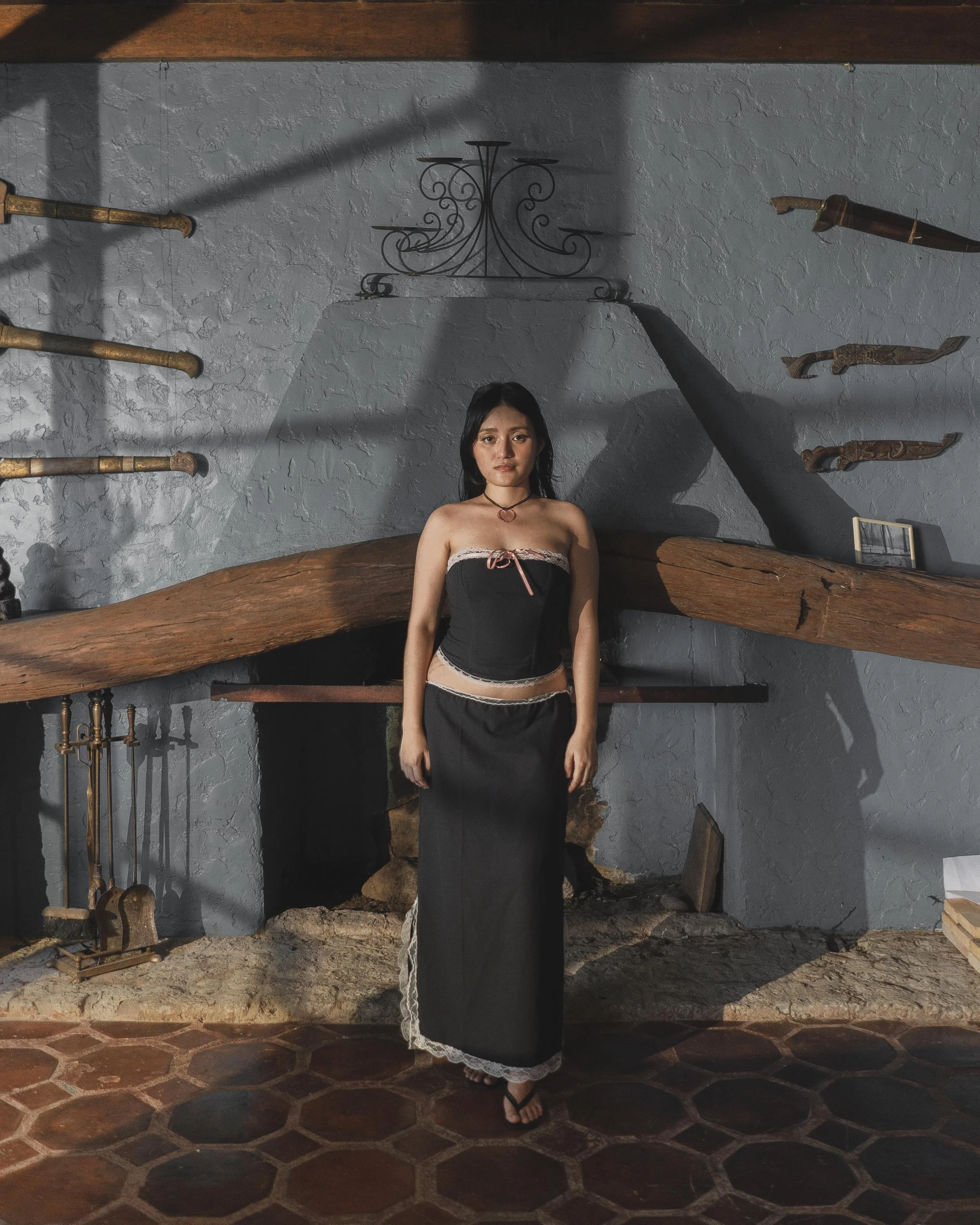 A young woman in a black strapless dress with lace trim standing in front of a rustic fireplace with wooden and metal decor, gray textured wall, and shadow patterns.