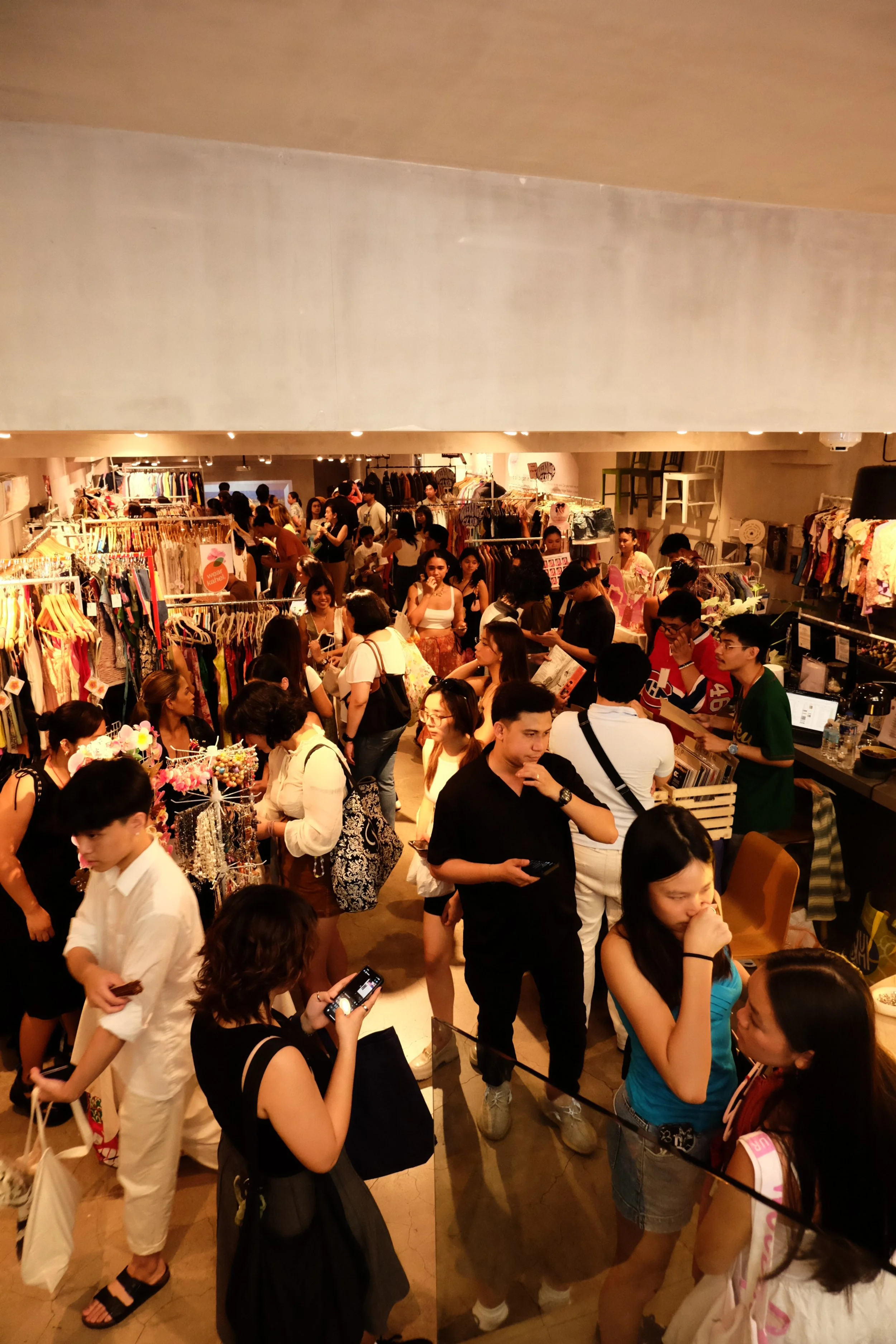Crowded clothing store with shoppers browsing and trying on clothes, displays of dresses and accessories, and a cashier counter in the background.