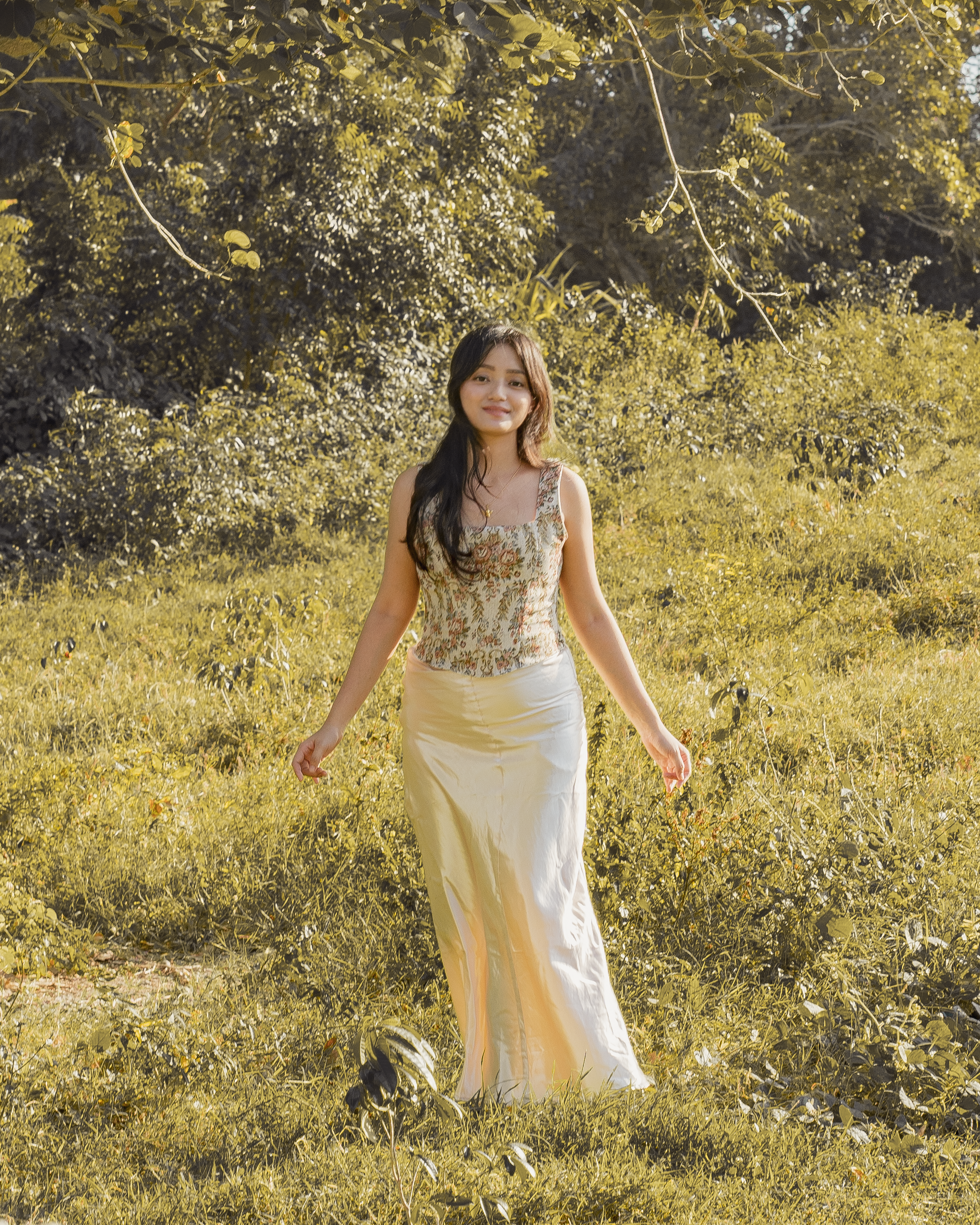 A young woman walking through a grassy field with trees in the background, wearing a sleeveless patterned top and a long cream skirt, smiling.