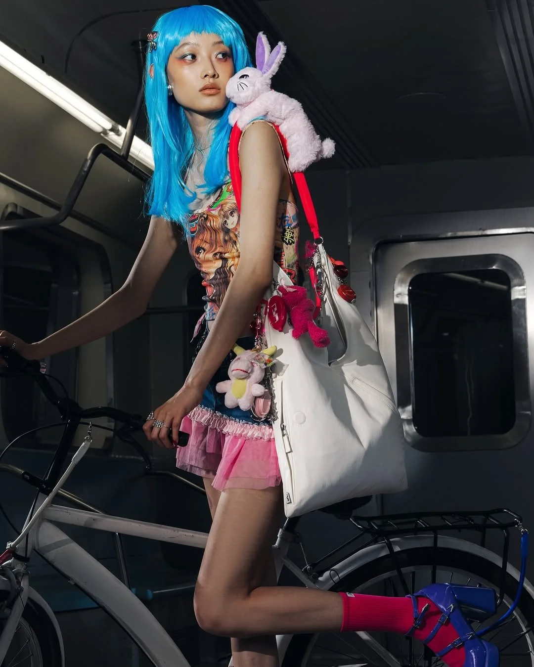 A young woman with bright blue hair riding a bicycle inside a subway train, carrying plush toys and a large white bag, wearing colorful clothing and high heels.