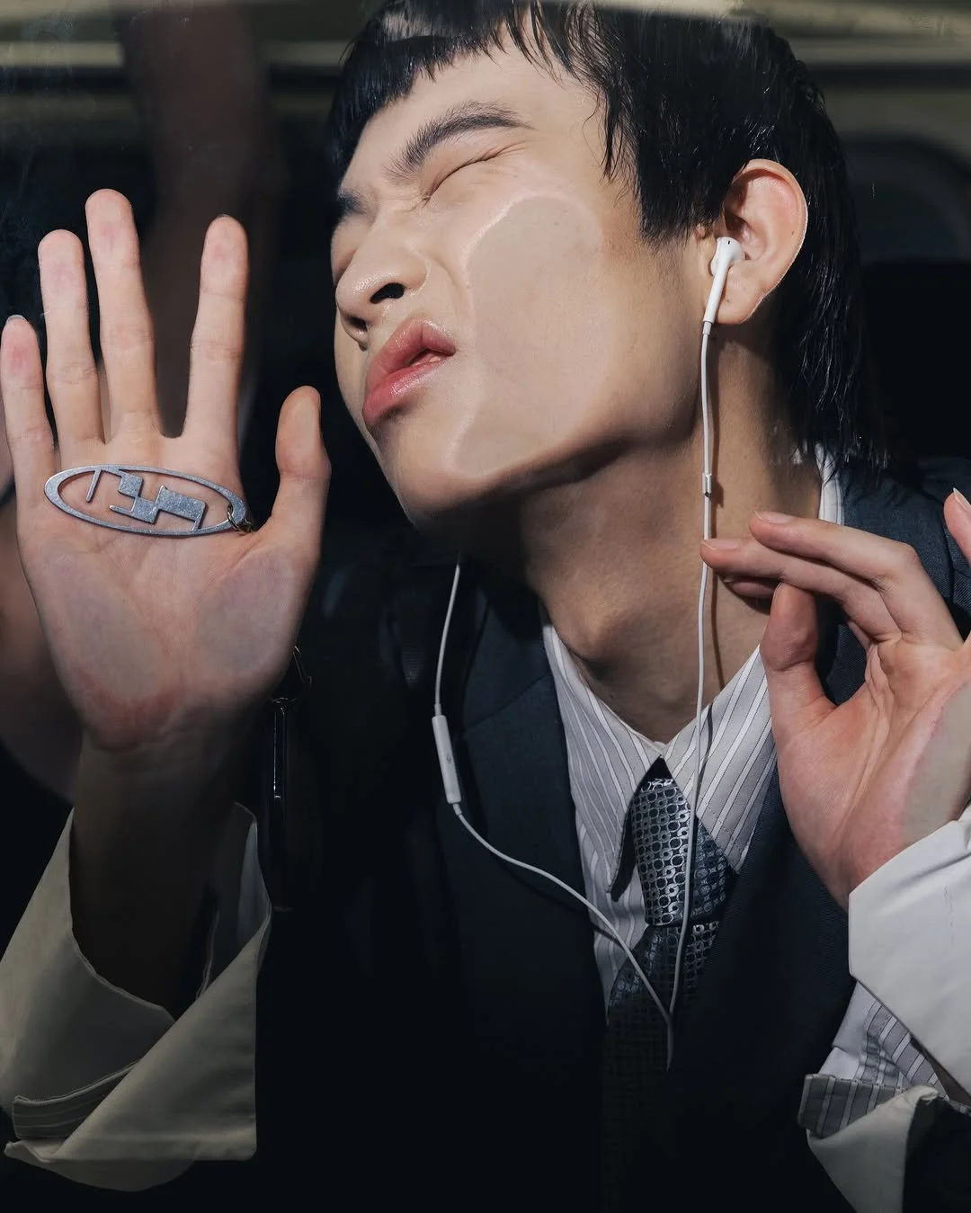 A person with black hair wearing a suit and earphones, eyes closed, appears to be enjoying music, with one hand raised and a silver ring on their finger.