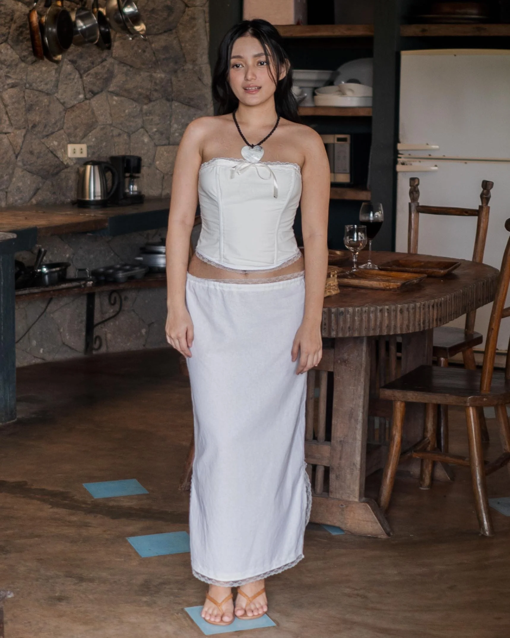 Young woman standing in a rustic kitchen, wearing a white strapless top and a long white skirt, with dark hair and a necklace with a large heart-shaped pendant.