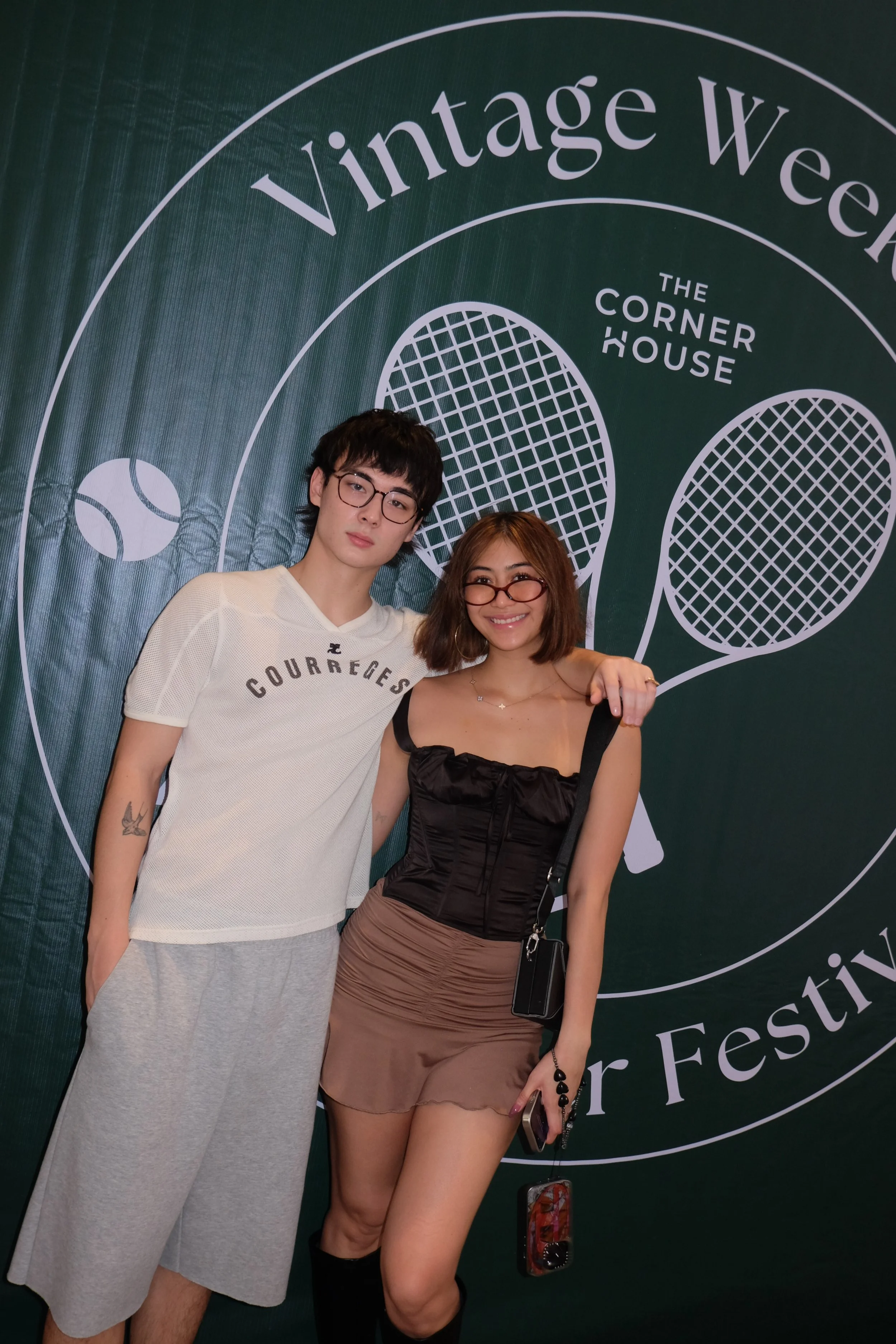 Two young people, a man and a woman, standing together at the 'Vintage Weekend' event at 'The Corner House'. The man has black hair, wears glasses and a casual white t-shirt with gray shorts. The woman has brown hair, also wears glasses, and is dressed in a black top and a brown skirt, holding a phone with a floral case. Behind them is a dark green backdrop with white text and graphics related to the event.