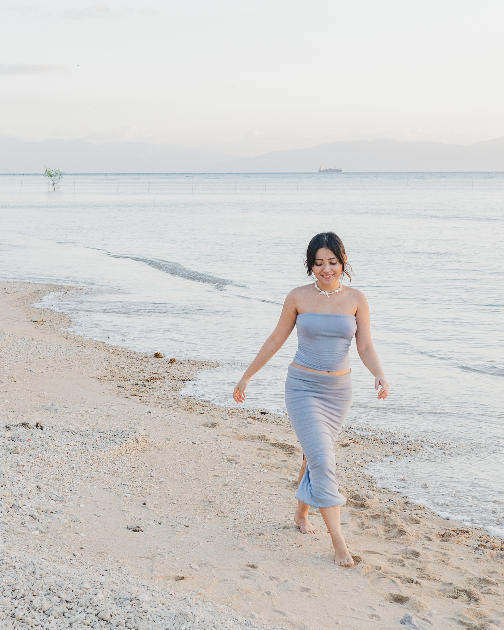 A woman in a light blue strapless dress and shell necklace walking barefoot on a sandy beach by the ocean.
