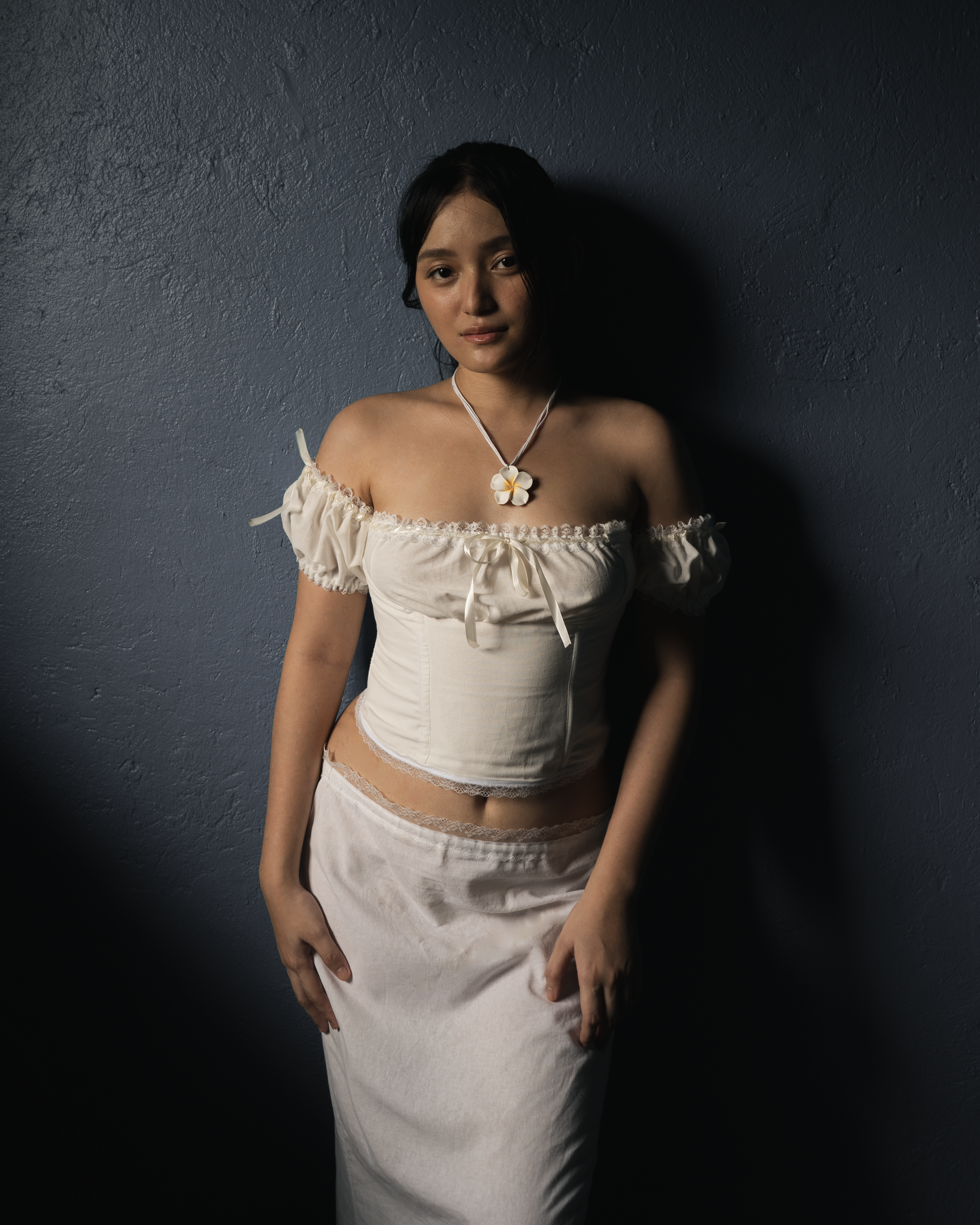 Young woman standing against a dark gray textured wall, wearing an off-the-shoulder white top, a long white skirt, and a floral pendant necklace.