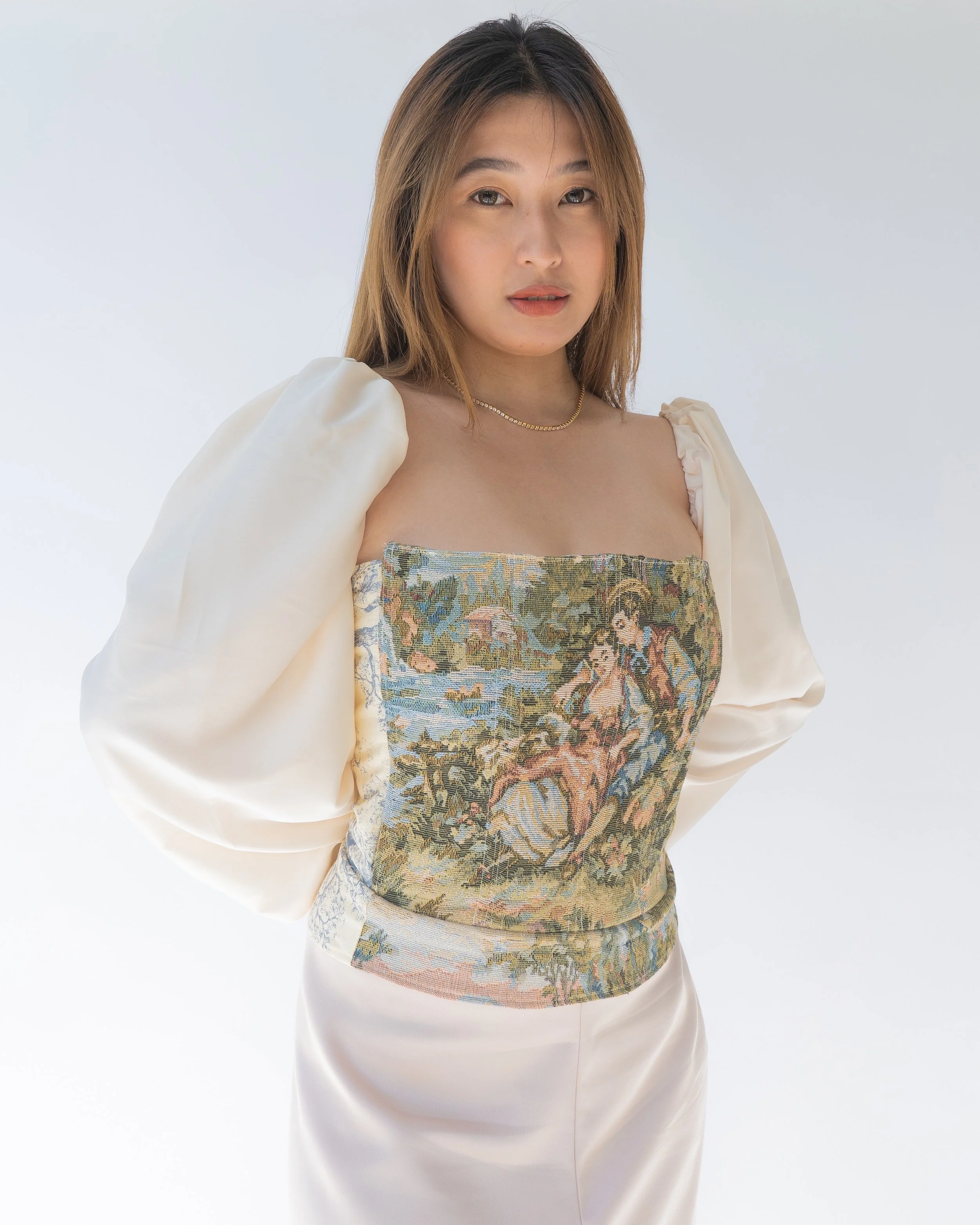 A young woman with light brown hair wearing a fashionable beige outfit with a tapestry-style top featuring a scene with a nude woman in nature