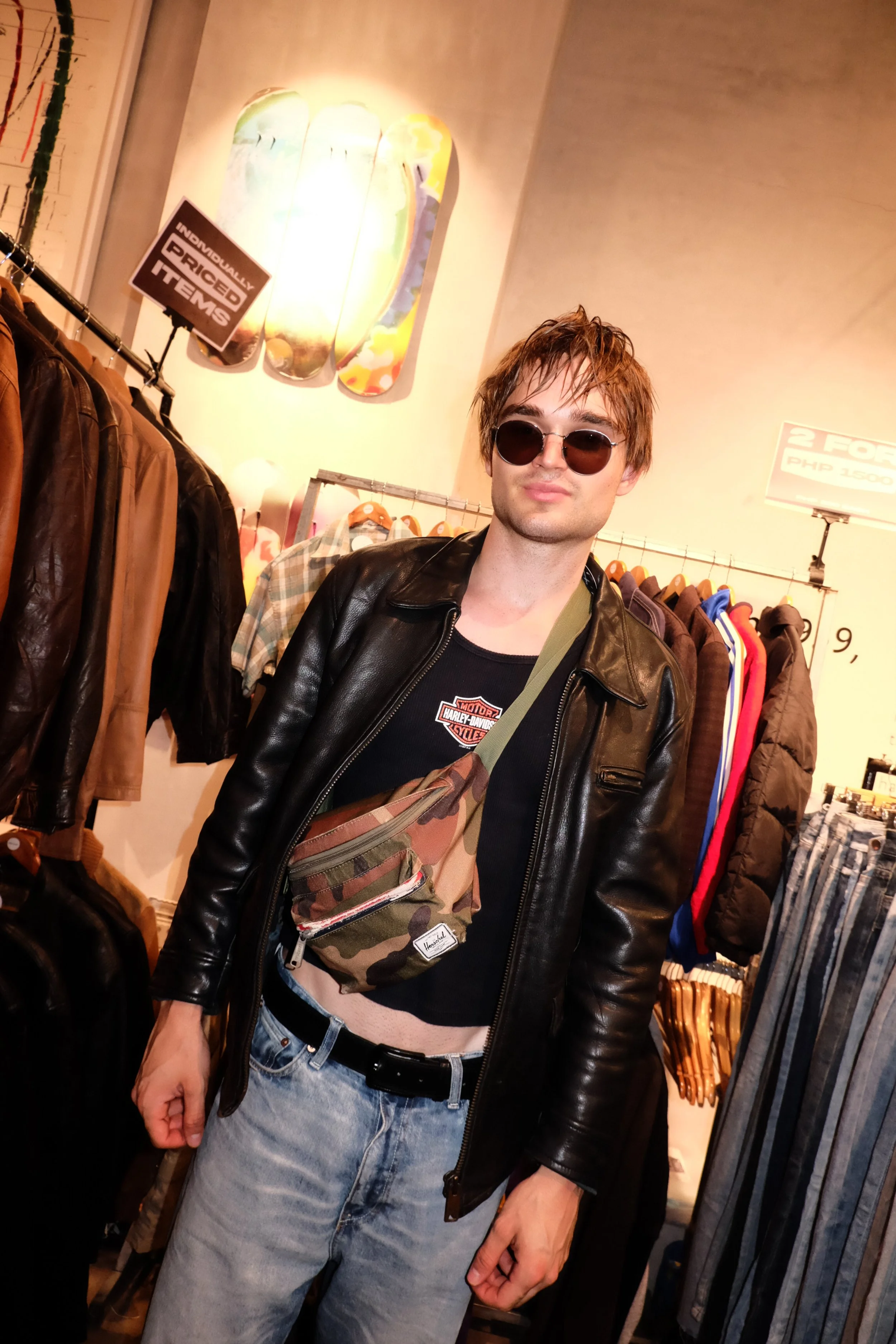 Young man with brown hair wearing sunglasses, a black leather jacket, black Harley Davidson t-shirt, light blue jeans, and camouflage crossbody bag shopping in a clothing store.