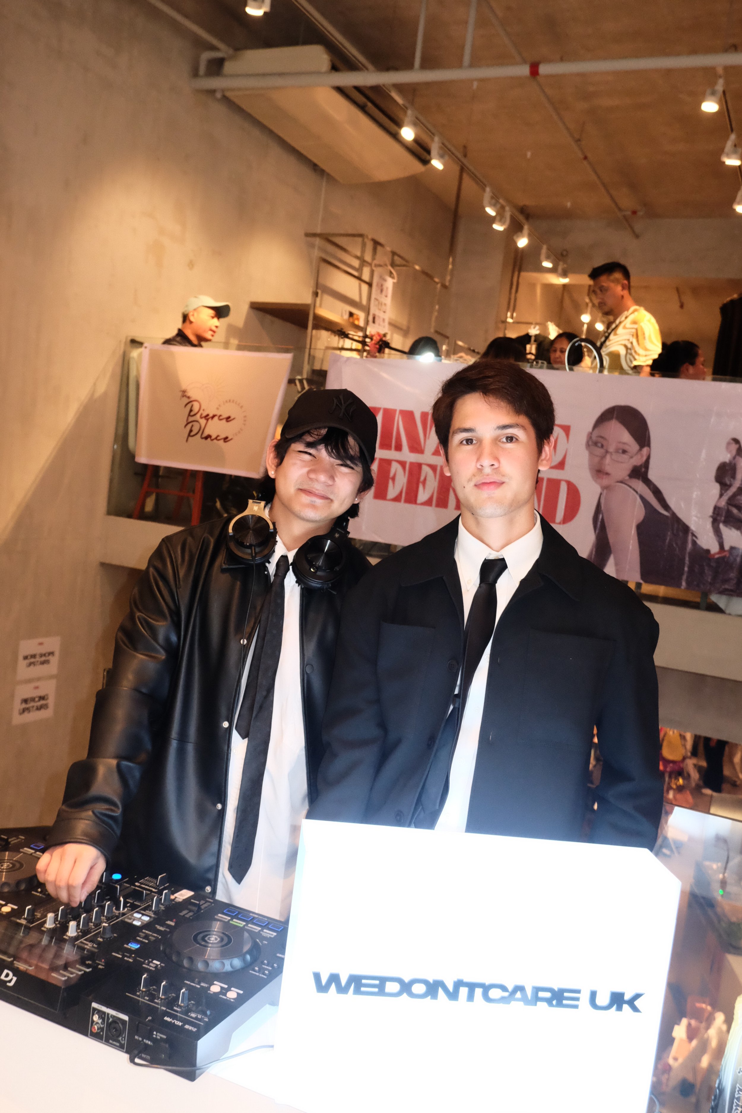 Two young men in black and white suits, one wearing a leather jacket and baseball cap and the other with short dark hair, standing behind DJ equipment at an event, with a lit sign that reads "WEDONTCARE UK" in front of them.