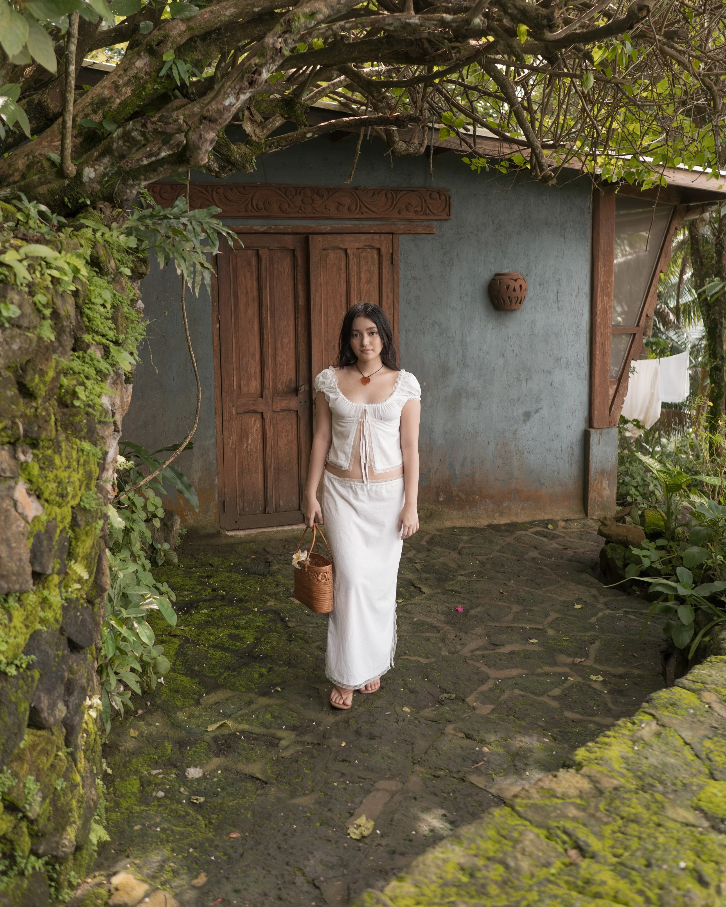 A young woman in a white outfit carries a woven bag and stands in front of a rustic house surrounded by greenery. The house has a gray wall, wooden door, and a small wall-mounted pot. There are trees and plants around, with laundry hanging outside.