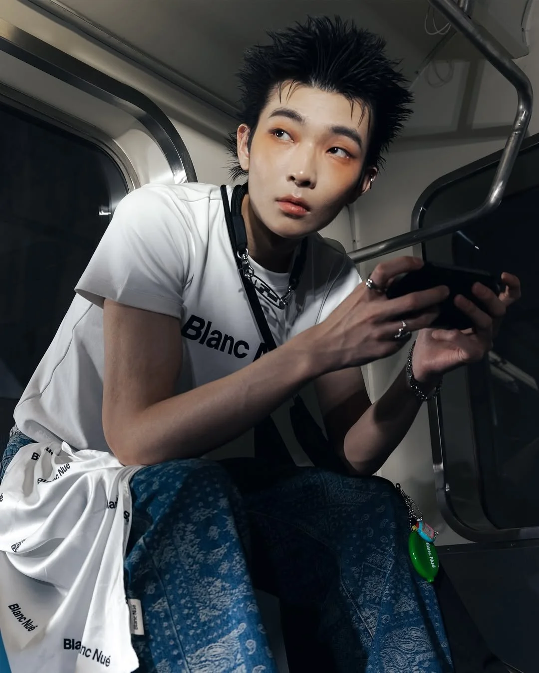 A young person with spiky black hair, wearing a white T-shirt and blue patterned pants, is sitting on a train and looking to the side while holding a smartphone.