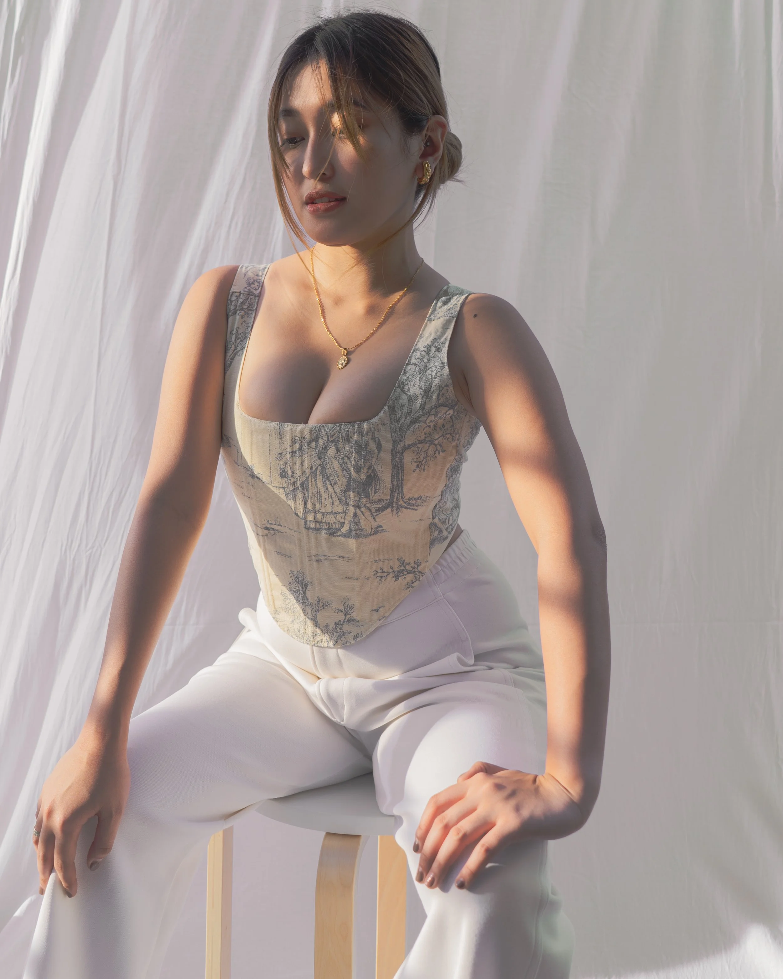 A woman with short hair sitting on a wooden stool, wearing a patterned sleeveless top and white pants, with soft lighting and a white fabric background.