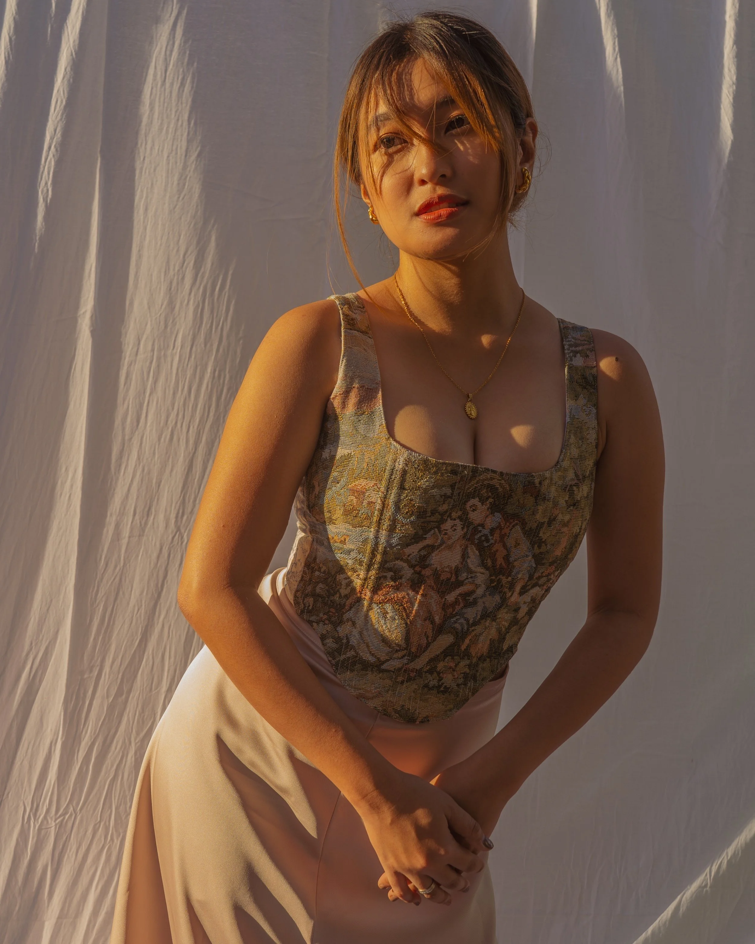 A woman standing in front of a white fabric backdrop, wearing a sleeveless floral top and a beige skirt, with sunlight casting shadows on her face and body.