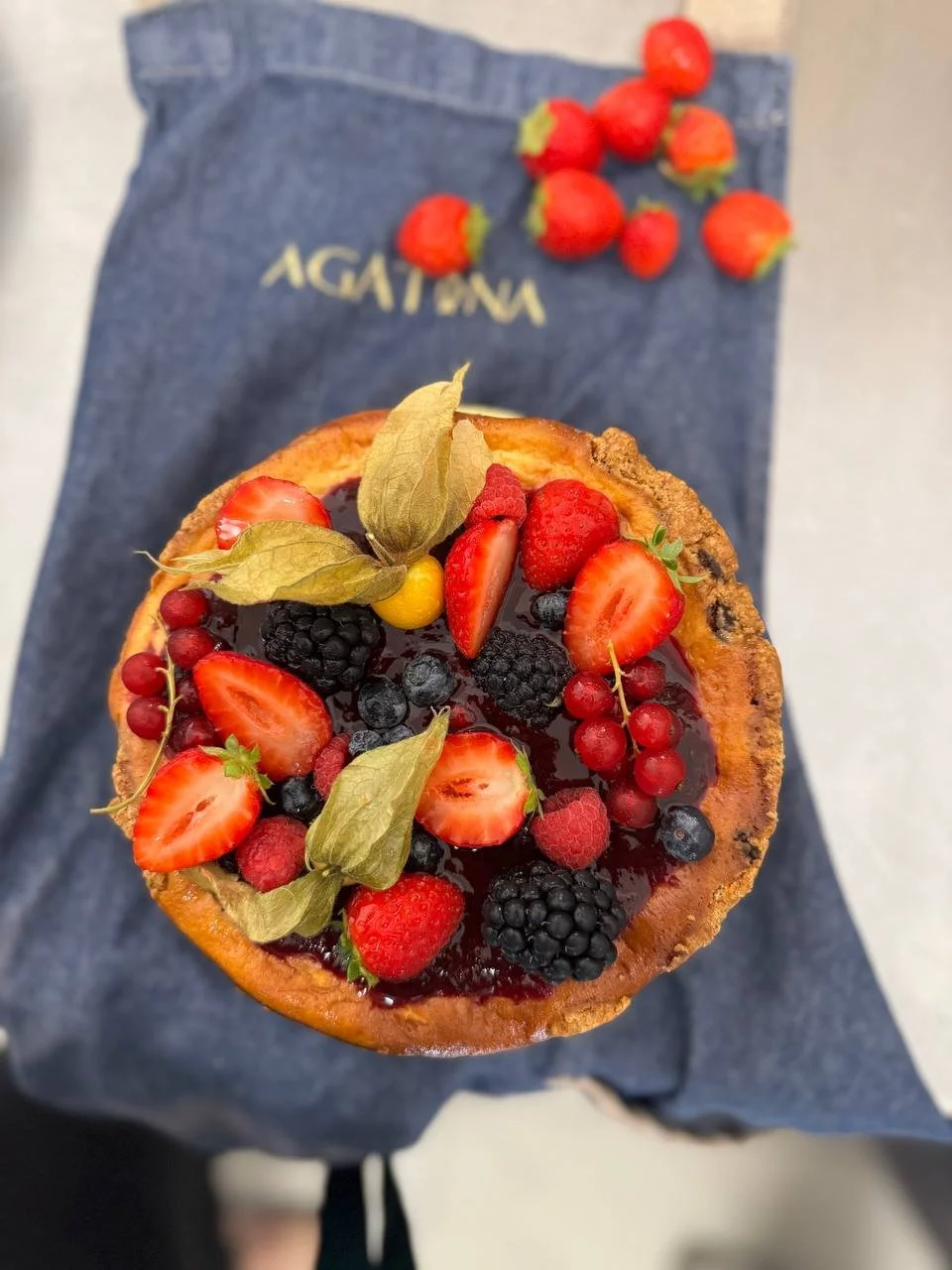 Torta con crema e frutti di bosco, decorata con fragole, lamponi, mirtilli, ribes, uva e foglie di basilico, posizionata su un lenzuolo blu.