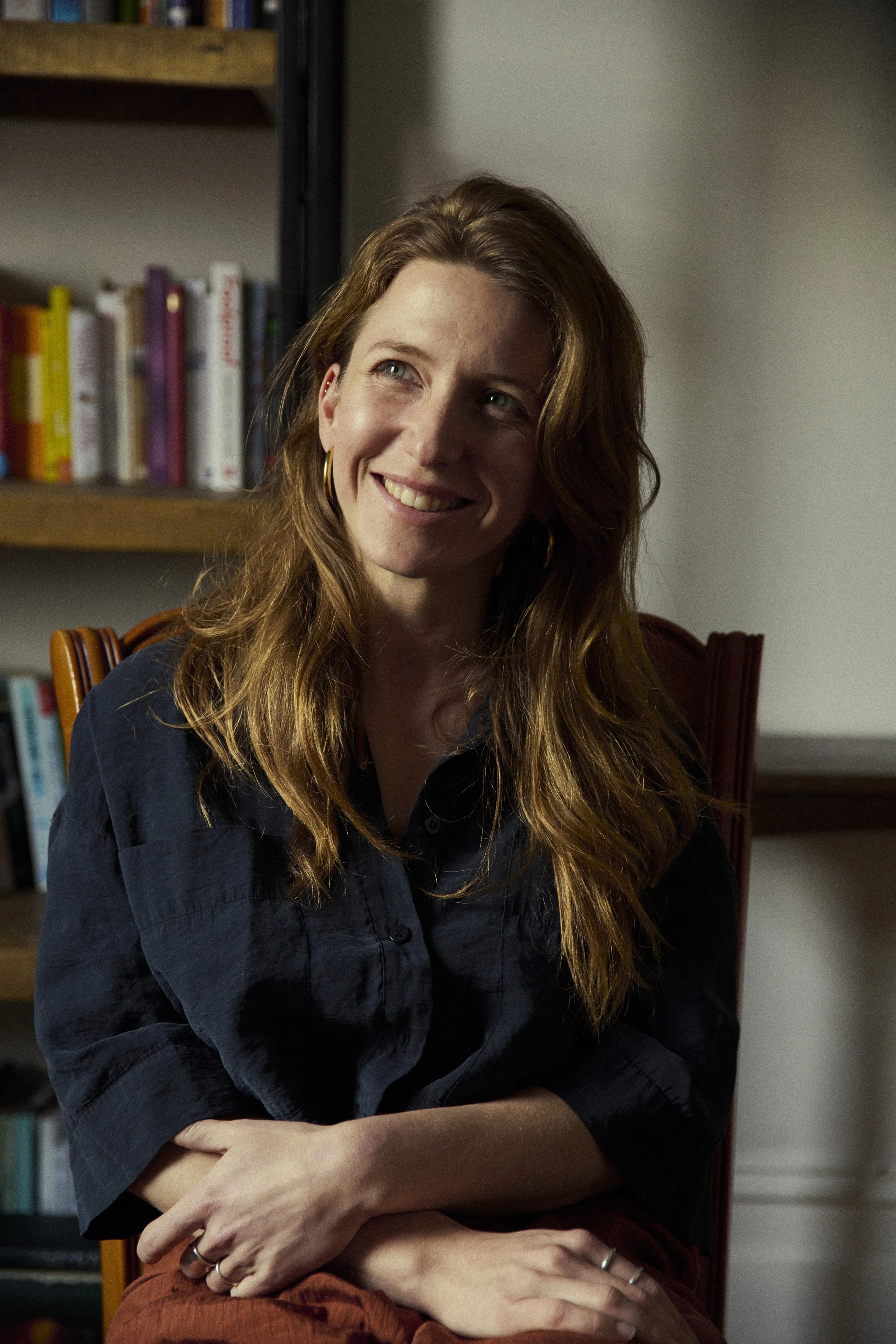 A woman with long, wavy red hair, wearing a black shirt and gold hoop earrings, sitting on a wooden chair with a slight smile, in a room with a bookshelf in the background.