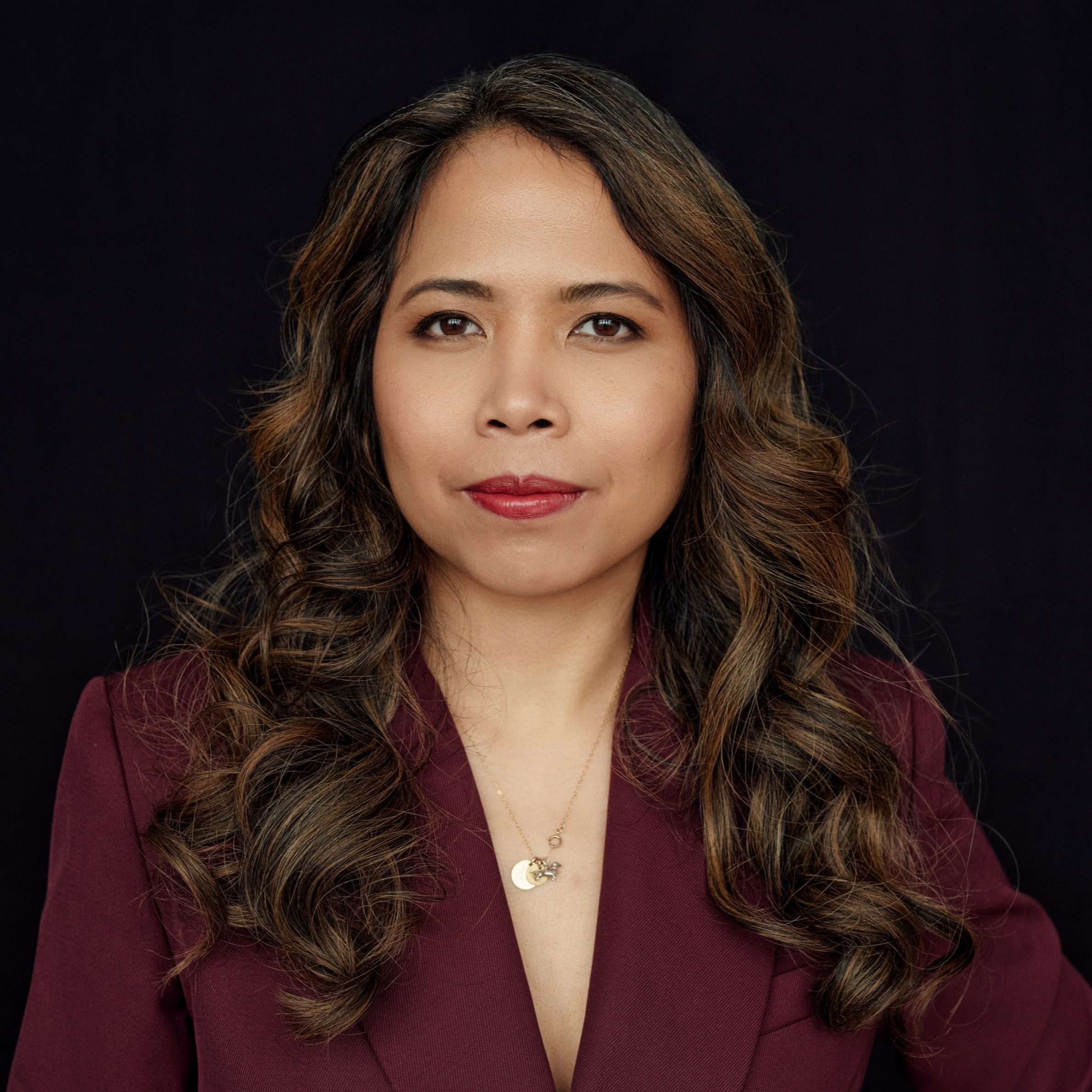 Professional headshot of a woman with long wavy brown hair wearing a burgundy blazer against a black background