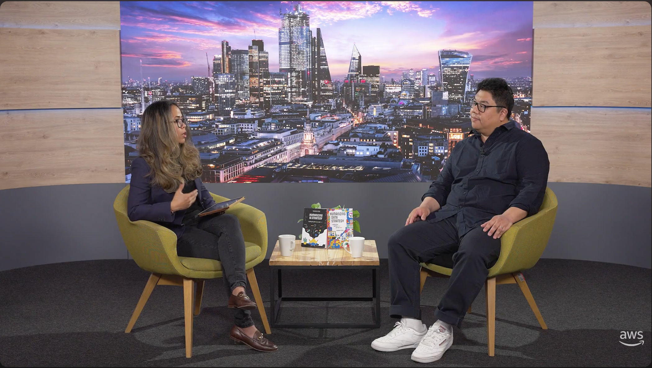 Two people sit in armchairs during an interview in front of a London skyline backdrop, with books and coffee on a table between them.