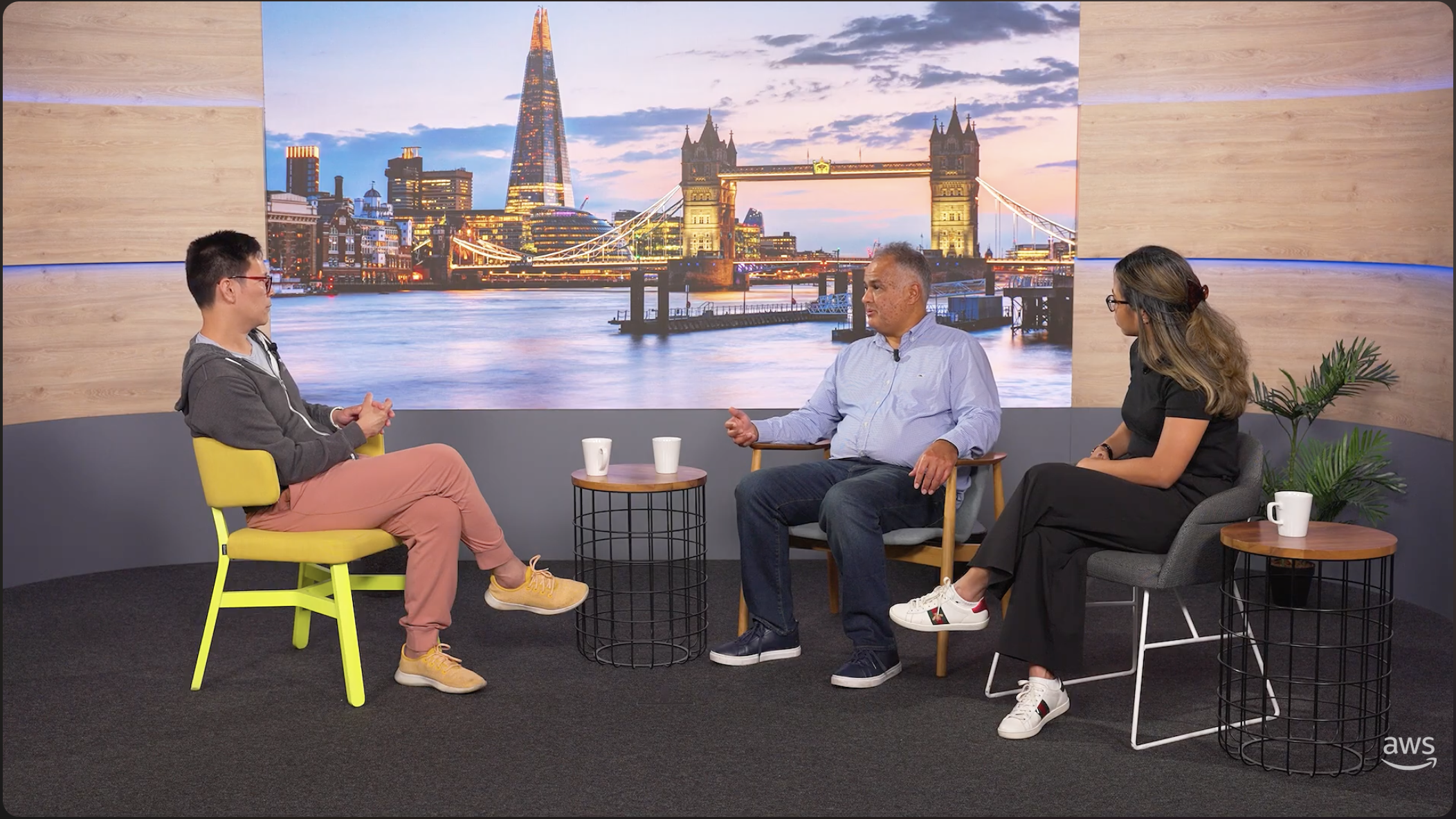 Three people discuss AWS technology in a studio with London’s Tower Bridge and Shard displayed behind them.