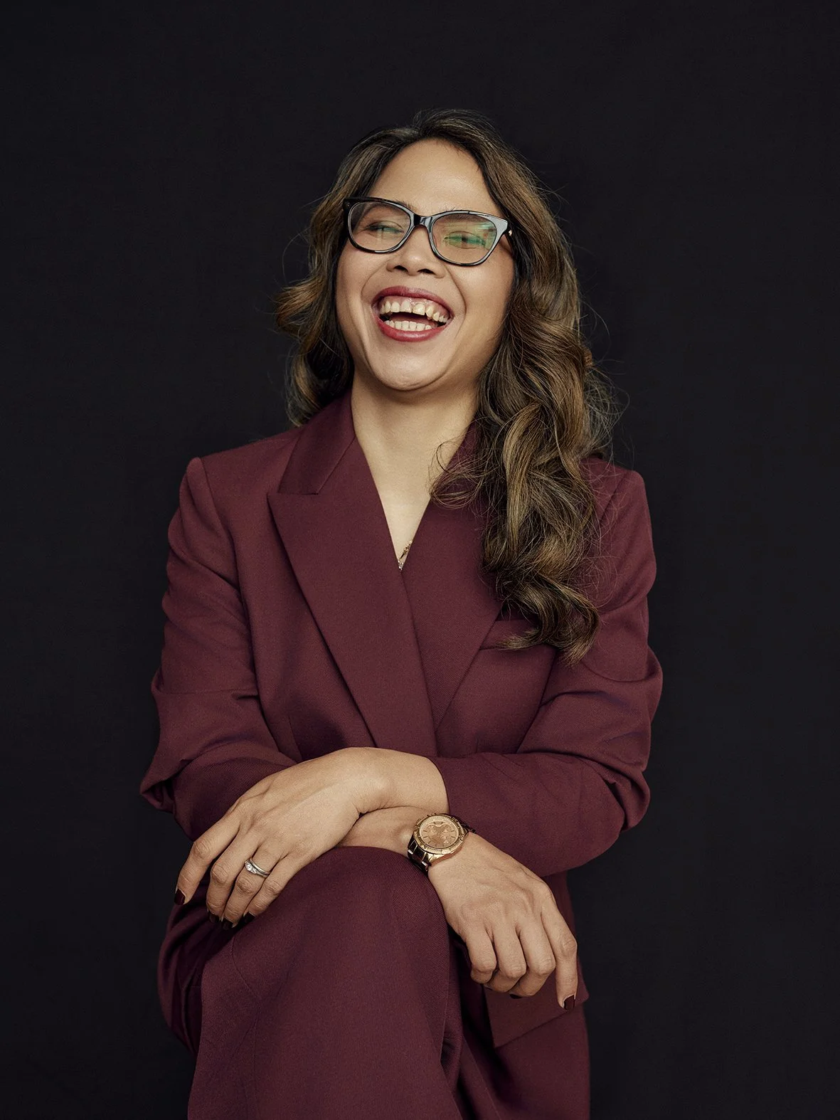 Smiling woman wearing glasses and a burgundy suit, seated against a dark background