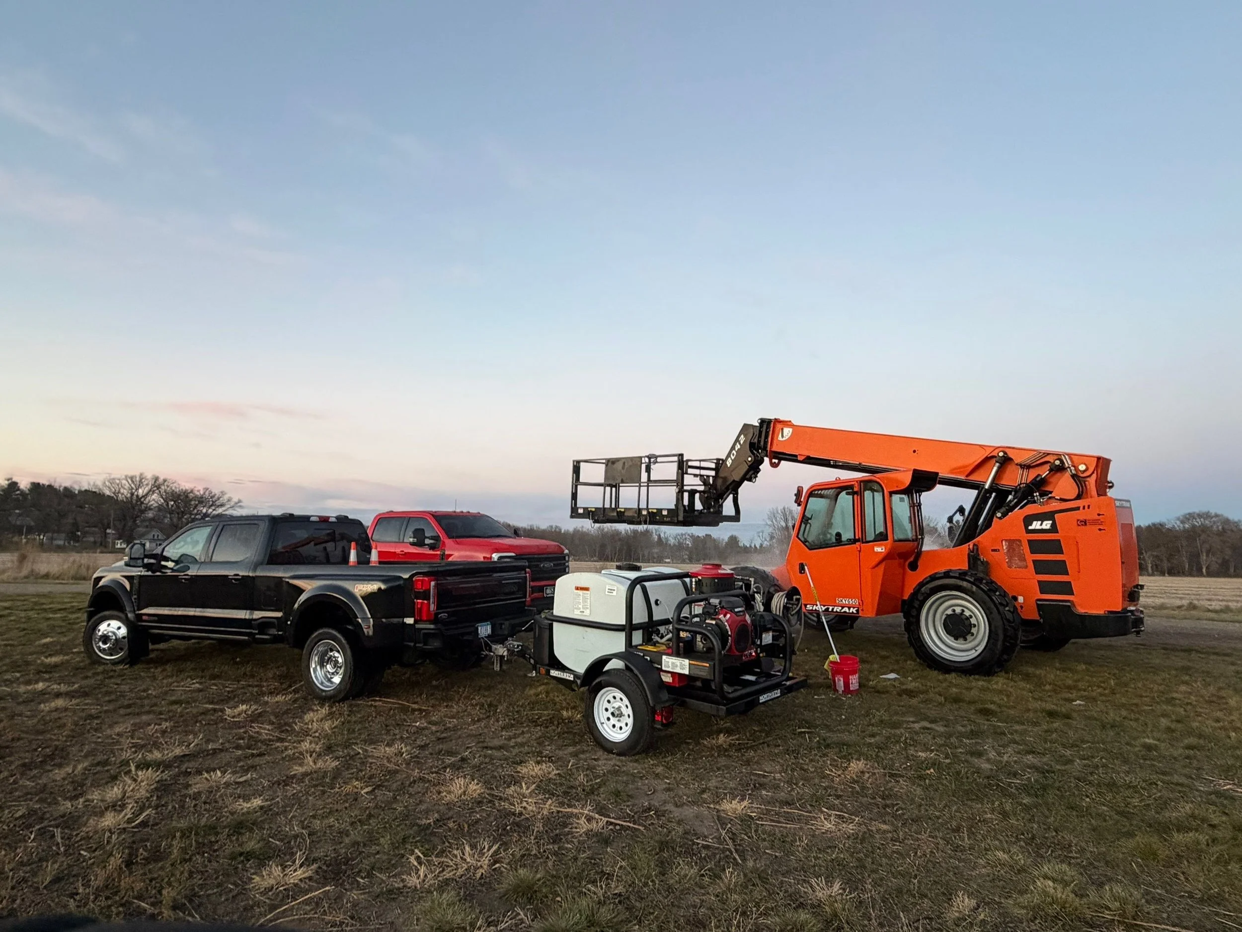 A collection of construction and farm equipment on a grassy field during sunset, including a black pickup truck, a red pickup truck, an orange lift, and a small mobile generator or pump.