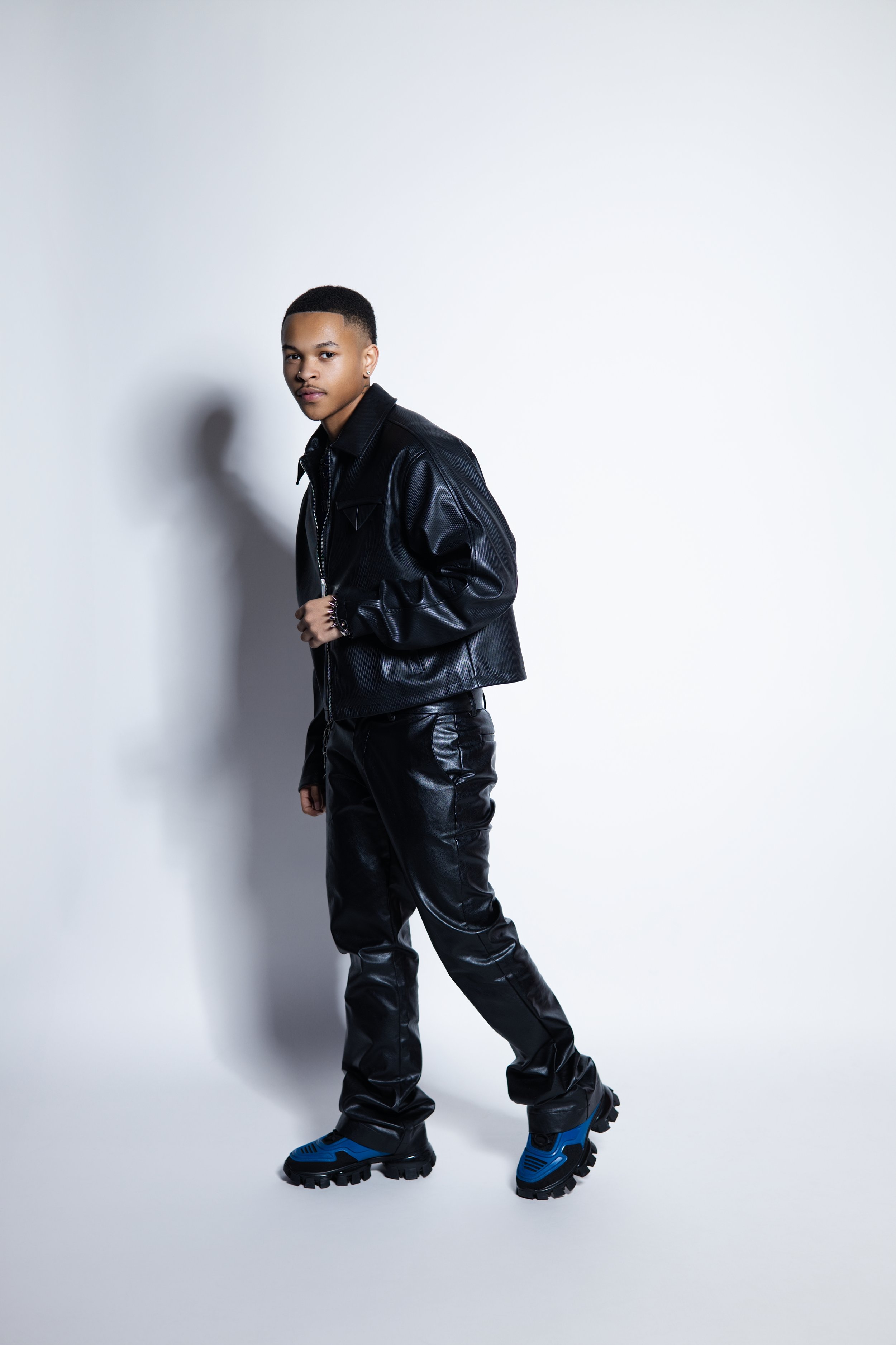 Young person wearing a black leather jacket, black leather pants, and blue and black hiking shoes, standing against a plain white background.