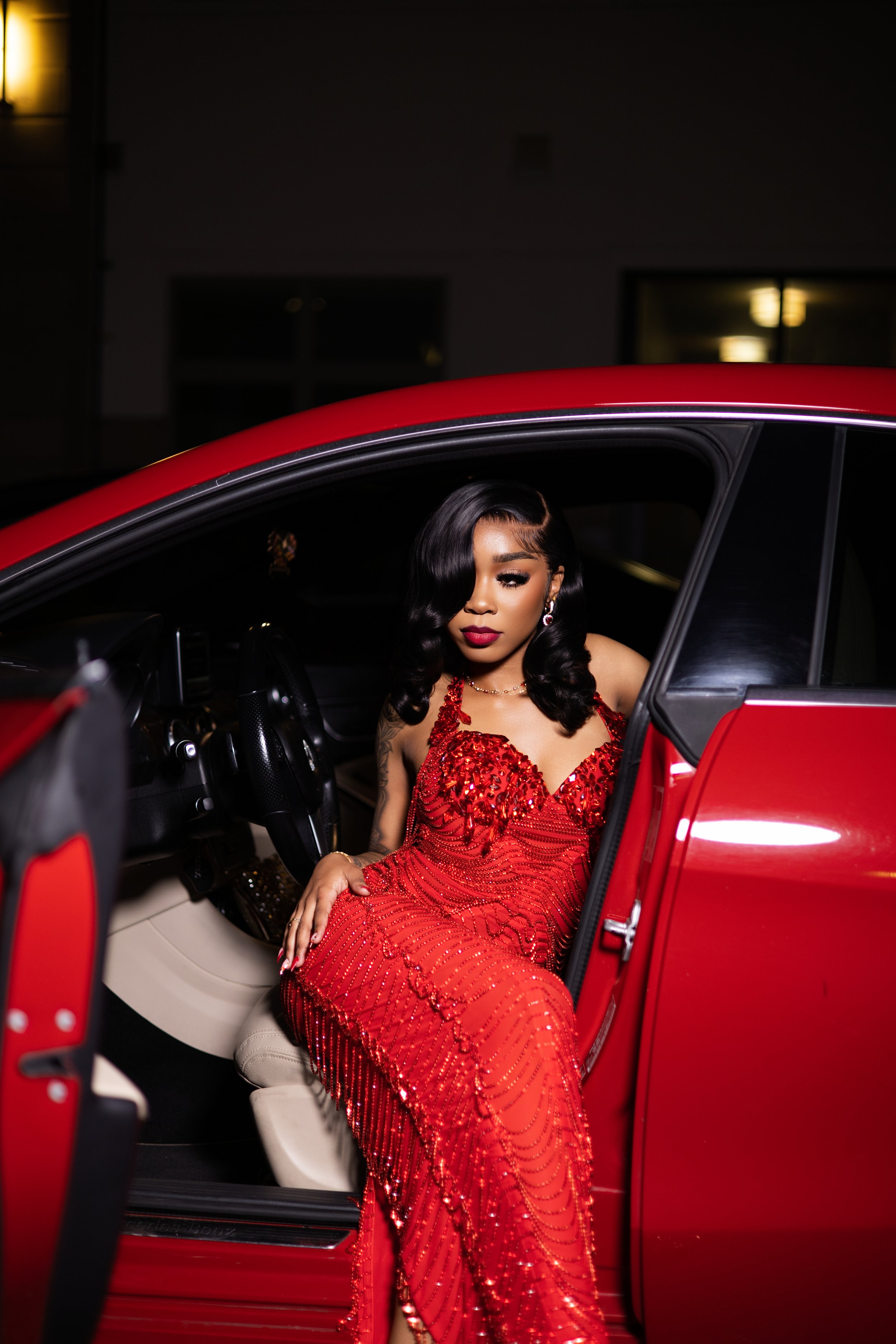 A woman in a red, sequined evening gown sitting inside a red luxury car with the door open at night.