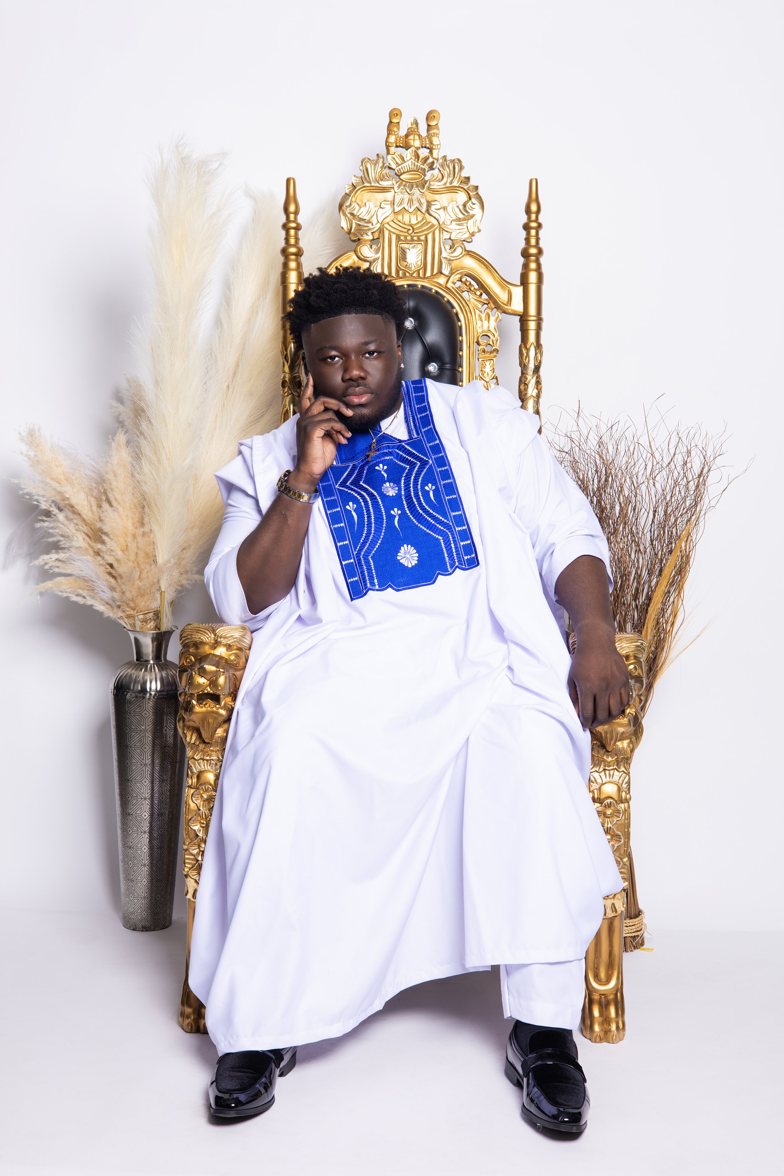 Young man sitting on a golden throne, wearing a white traditional outfit with blue embroidery, and dark shoes. Behind him are decorative vases with pampas grass and dried branches.