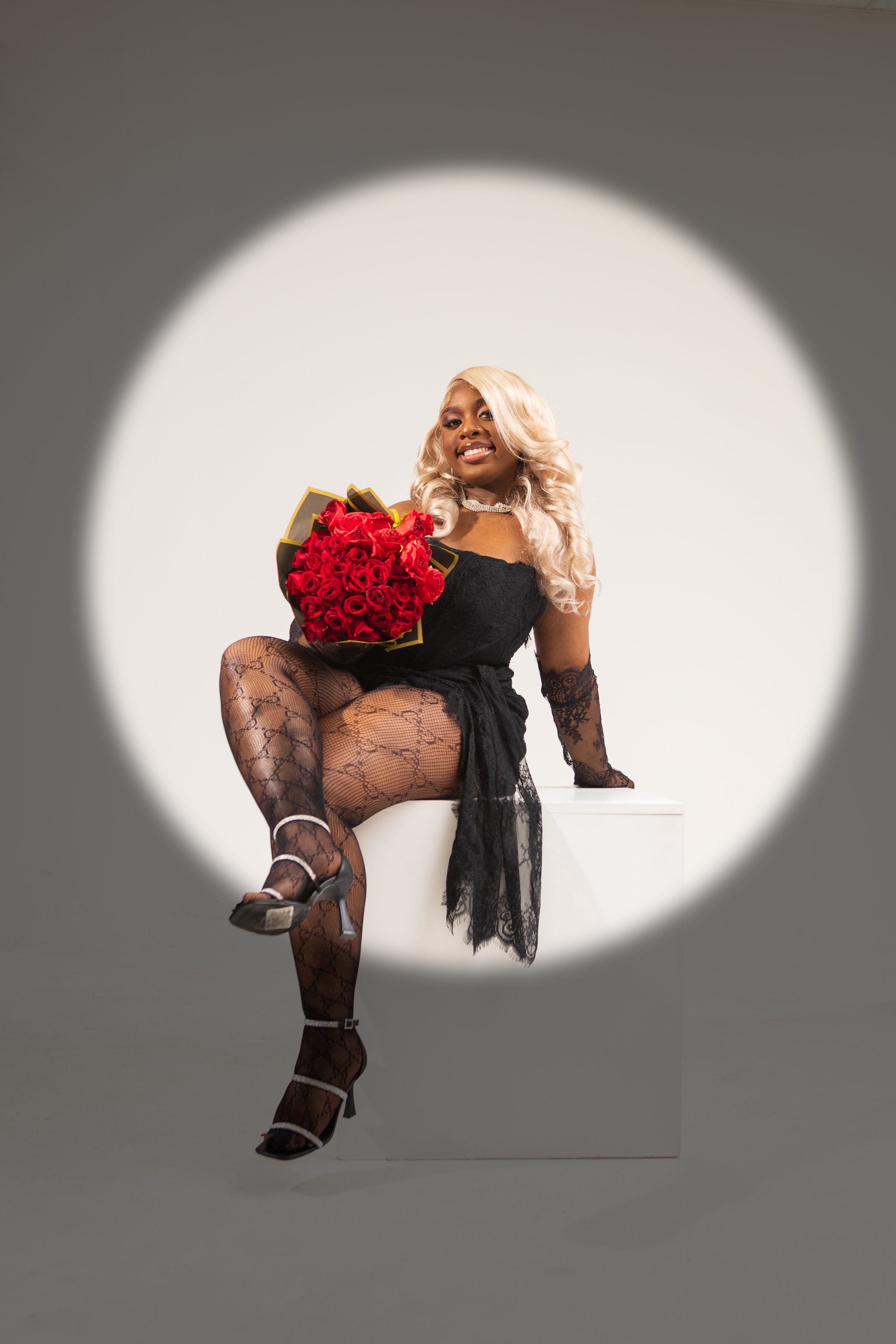 A woman with blonde curly hair sitting on a white block, holding a large bouquet of red roses, wearing black lace clothing and high heels, smiling at the camera, framed by a circular spotlight on a plain background.