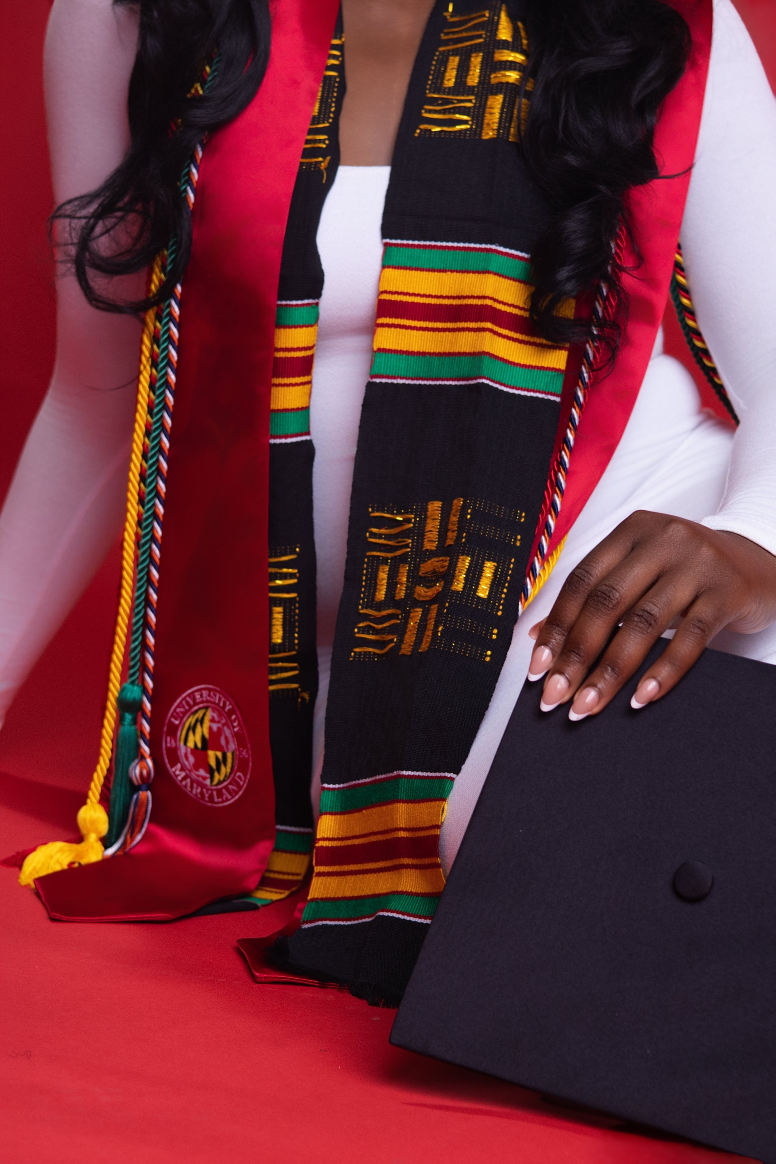 Close-up of a person in graduation attire, wearing a white long-sleeve shirt, with a black and colorful honor stole decorated with gold, red, green, and yellow stripes, and an academic cap on a red surface.