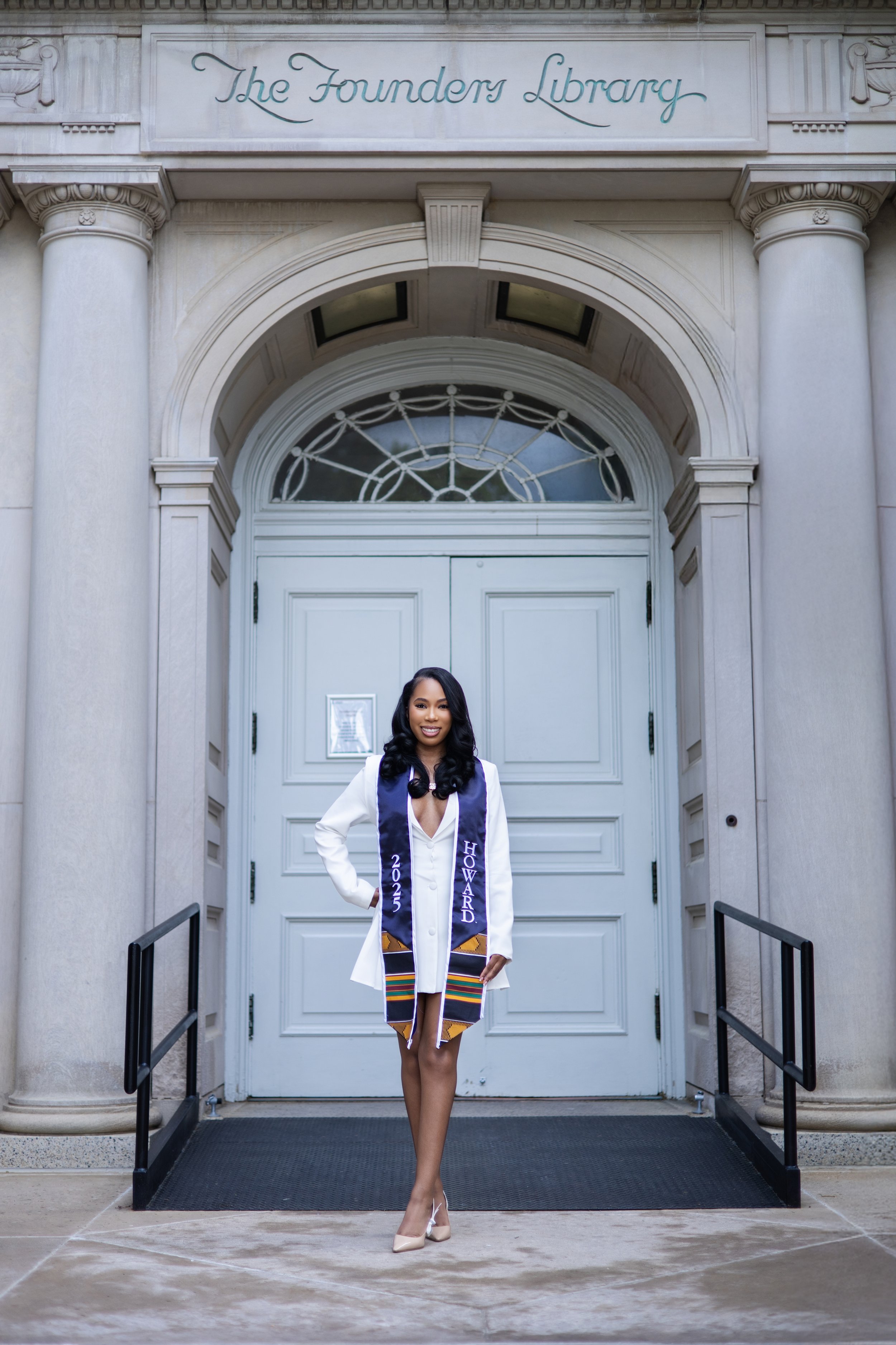 A woman in a white dress and heels standing in front of a courthouse door, wearing a graduation stole with the year 2025 and the name Howard.