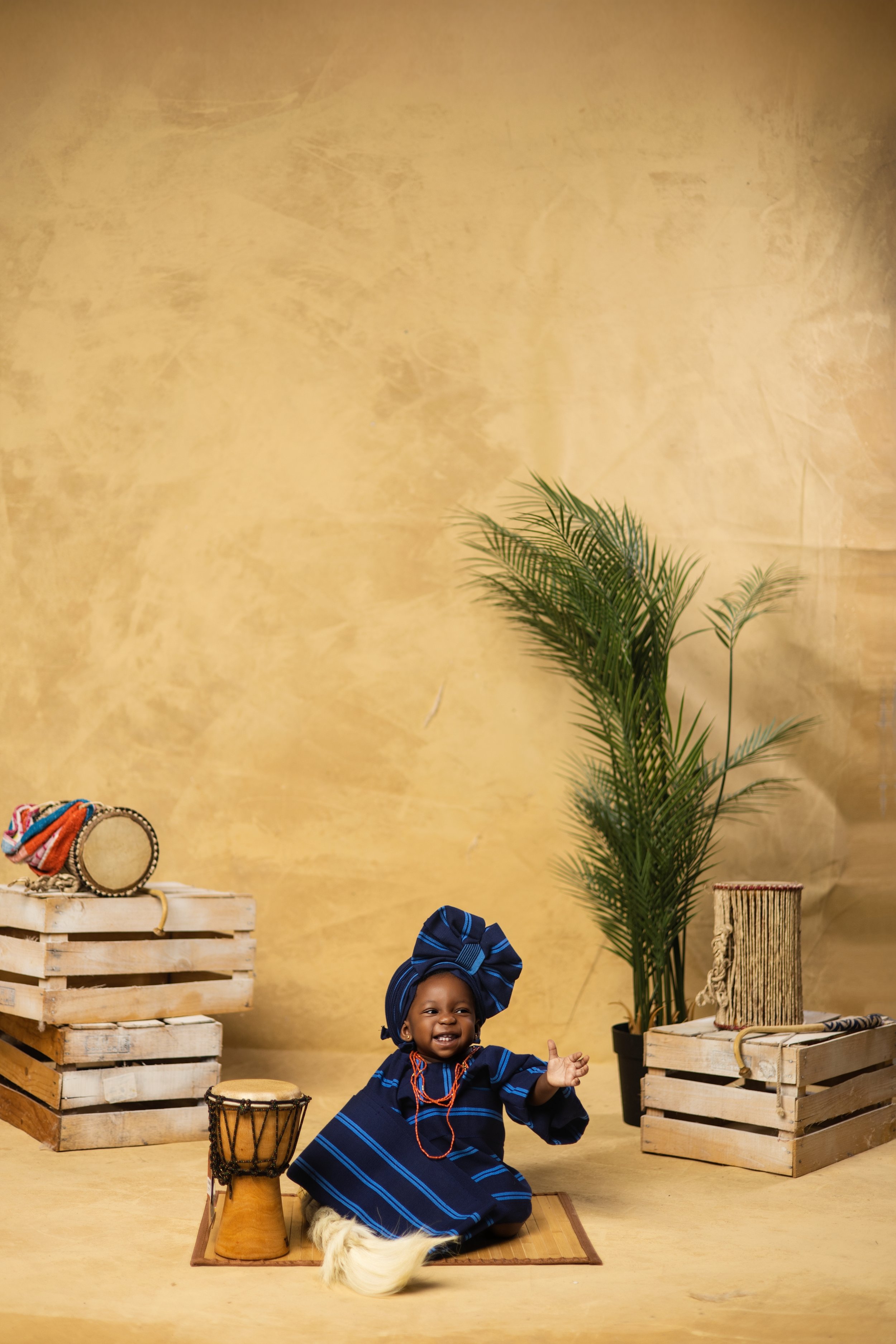 A smiling child dressed in traditional African attire with a large headwrap, sitting on a mat with drums and wooden crates around, and a potted plant in the background.